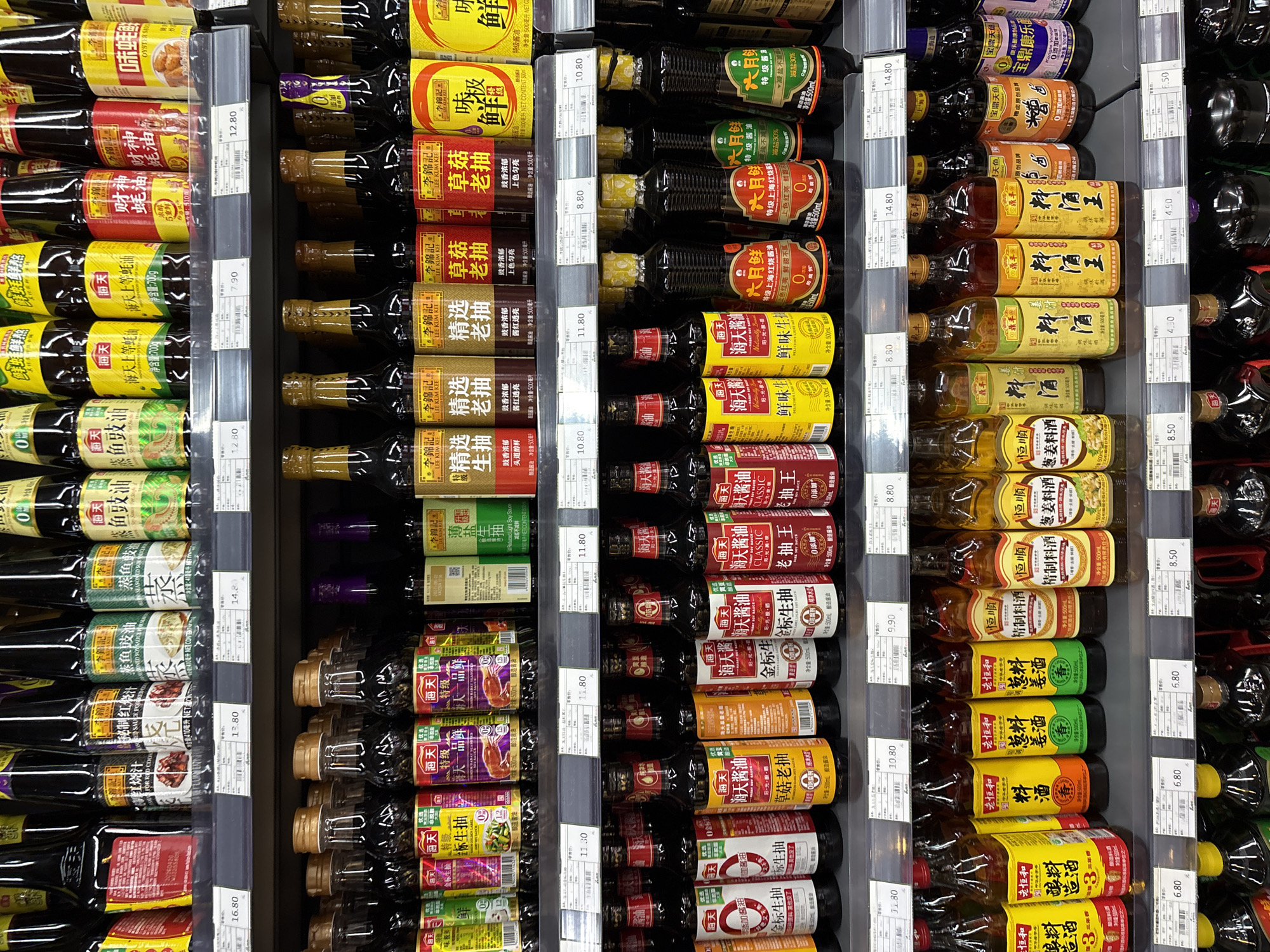 The image depicts a retail shelf in a supermarket located in Minhang District, China. The shelf is stocked with a variety of bottled condiments, primarily soy sauce and cooking wine. The products are neatly arranged in rows, with labels in both Chinese and English. The lighting is bright and even, typical of a retail environment. There are no people visible in the frame. The overall scene suggests a well-organized and stocked supermarket aisle.