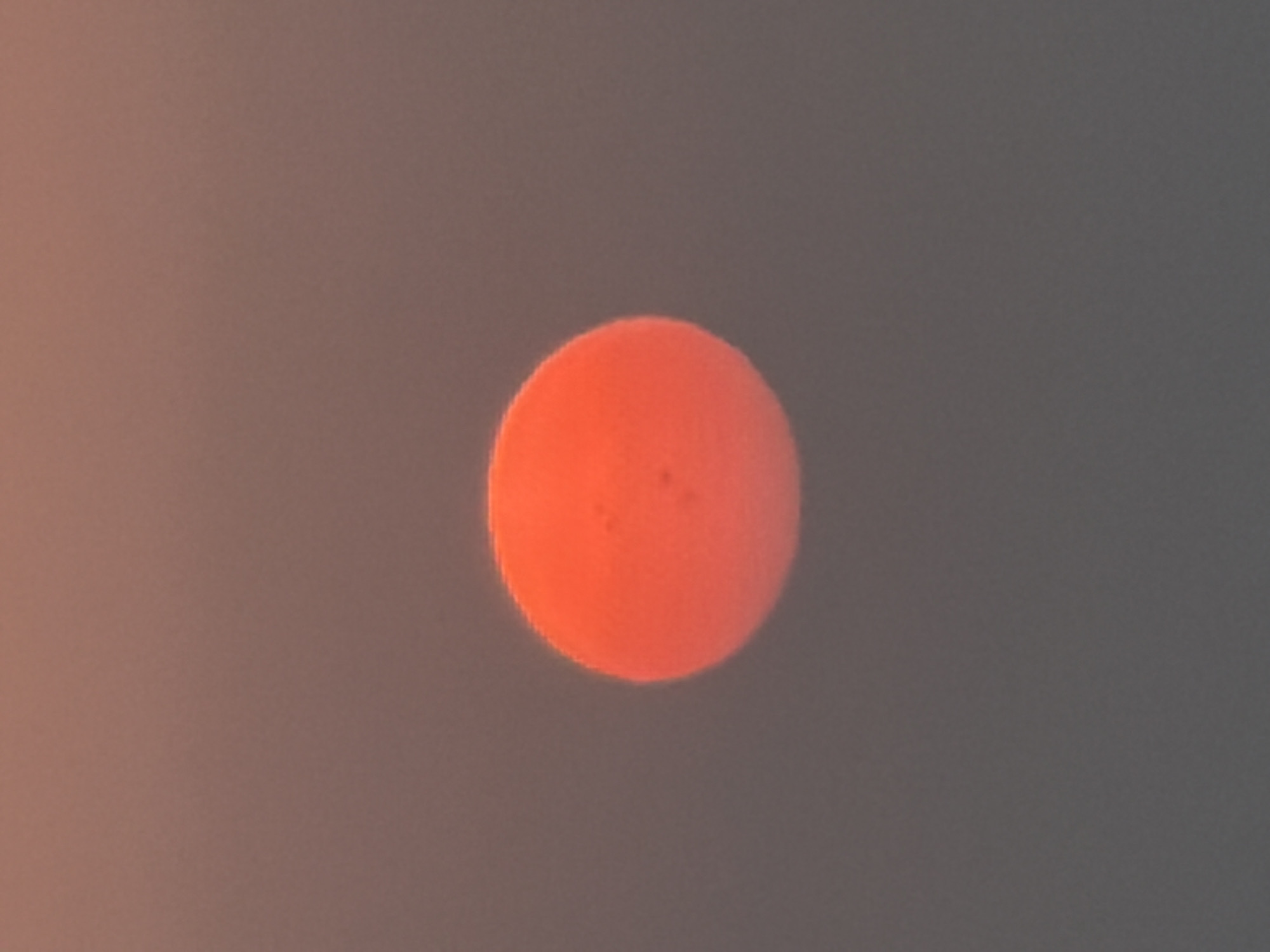 A low-angle, full-frame shot captures a bright orange, spherical sun against a hazy, pale orange sky, suggesting it is either sunrise or sunset. The sun is positioned slightly left of the center of the frame. The image is slightly out of focus, but appears to show some darker, subtle markings on the surface of the sun, possibly sunspots or atmospheric distortions. The environment is not discernable beyond the sky and the sun.