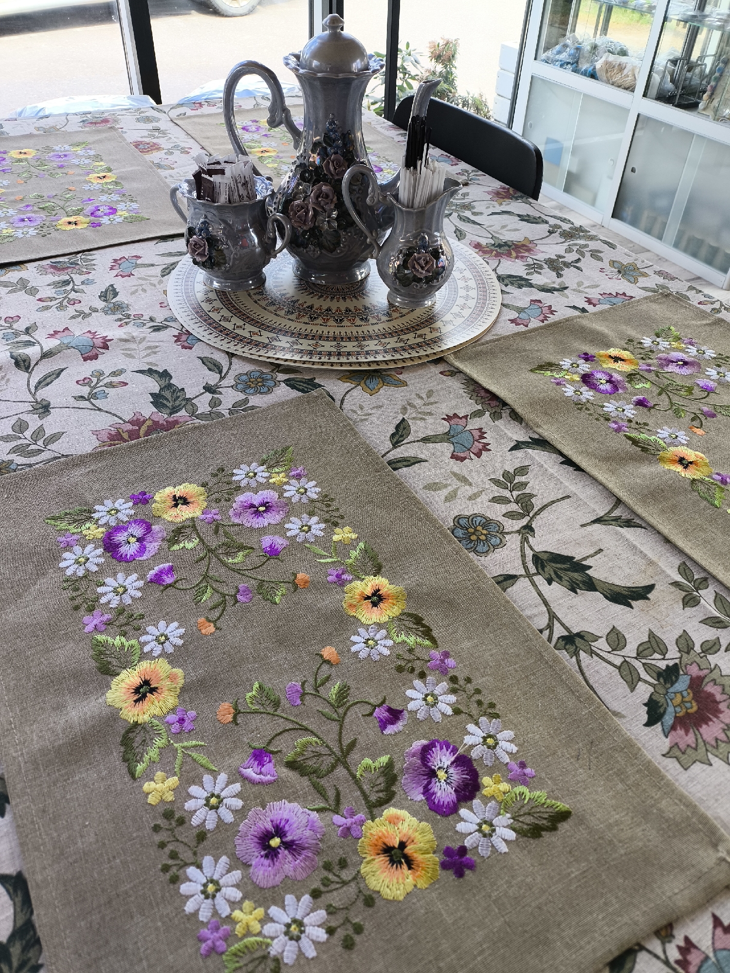 This image presents a beautifully arranged table setting, likely in a home or a small eatery, situated in Kluschino, Russia. The central focus is a rectangular table covered with a light-colored tablecloth adorned with a sprawling, ornate floral pattern featuring large stylized blooms and foliage in shades of blue, pink, and green.
On top of the tablecloth are several rectangular placemats or runners, made of a rustic, possibly linen-like, tan fabric. These are intricately hand-embroidered with vibrant, detailed floral designs. The embroidery showcases a rich assortment of flowers, including prominent yellow and purple pansies, delicate white and smaller yellow daisies, and tiny purple and orange blossoms, all interconnected by green stems and leaves. The floral arrangements form graceful, organic wreaths or garlands on each placemat.
At the center of the table, resting on a round decorative charger with intricate geometric patterns, is a matching three-piece ceramic set. This elegant set, in a muted grey color, includes a lidded teapot or coffee pot, a sugar bowl filled with individual sugar packets, and a creamer or pitcher containing stirrers or spoons. Each piece is embellished with raised, three-dimensional floral decorations in subtle, complementary colors.
The scene is illuminated by ample natural light entering through a large window on the left, indicating it is daytime. The view outside is indistinct but shows general outdoor light. In the background to the right, a white cabinet with glass-fronted doors, possibly storing various items or craft supplies, is visible, along with a portion of a black chair. The table is set for an activity such as a tea or coffee service. No people are present, and no discernible text is visible within the frame.