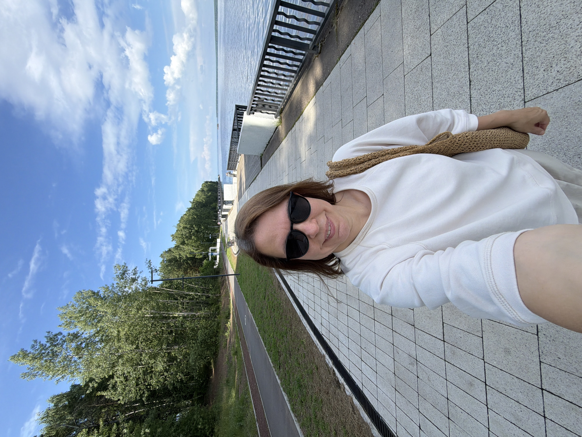 A woman with brown hair and sunglasses takes a selfie on a paved promenade. She is wearing a white crew neck sweatshirt and light-colored pants. A brown woven bag is slung over her right shoulder. Behind her, the promenade stretches along a body of water, with a black metal railing on the right. On the left, there are trees and a walking/biking path. The sky is blue with scattered white clouds. This scene is located in Chaikovsky, Russia.