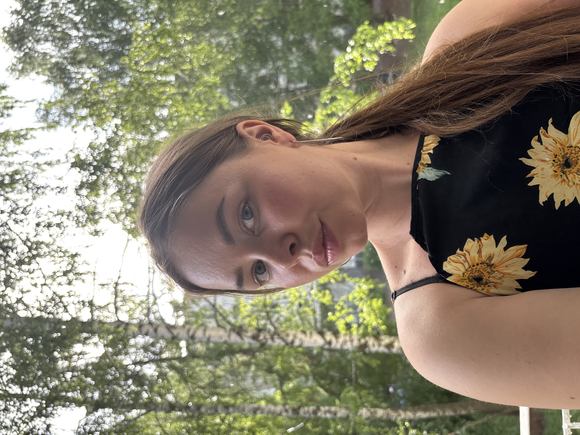 A person with long brown hair, wearing a black top with sunflower patterns, is depicted in an outdoor setting. The individual is angled slightly, with their face and upper body visible. Green foliage and tree branches are discernible in the background, suggesting a park or garden setting. The trees appear to be silver birch trees, common in Izhevsk, Russia. The background is blurred, emphasizing the subject.