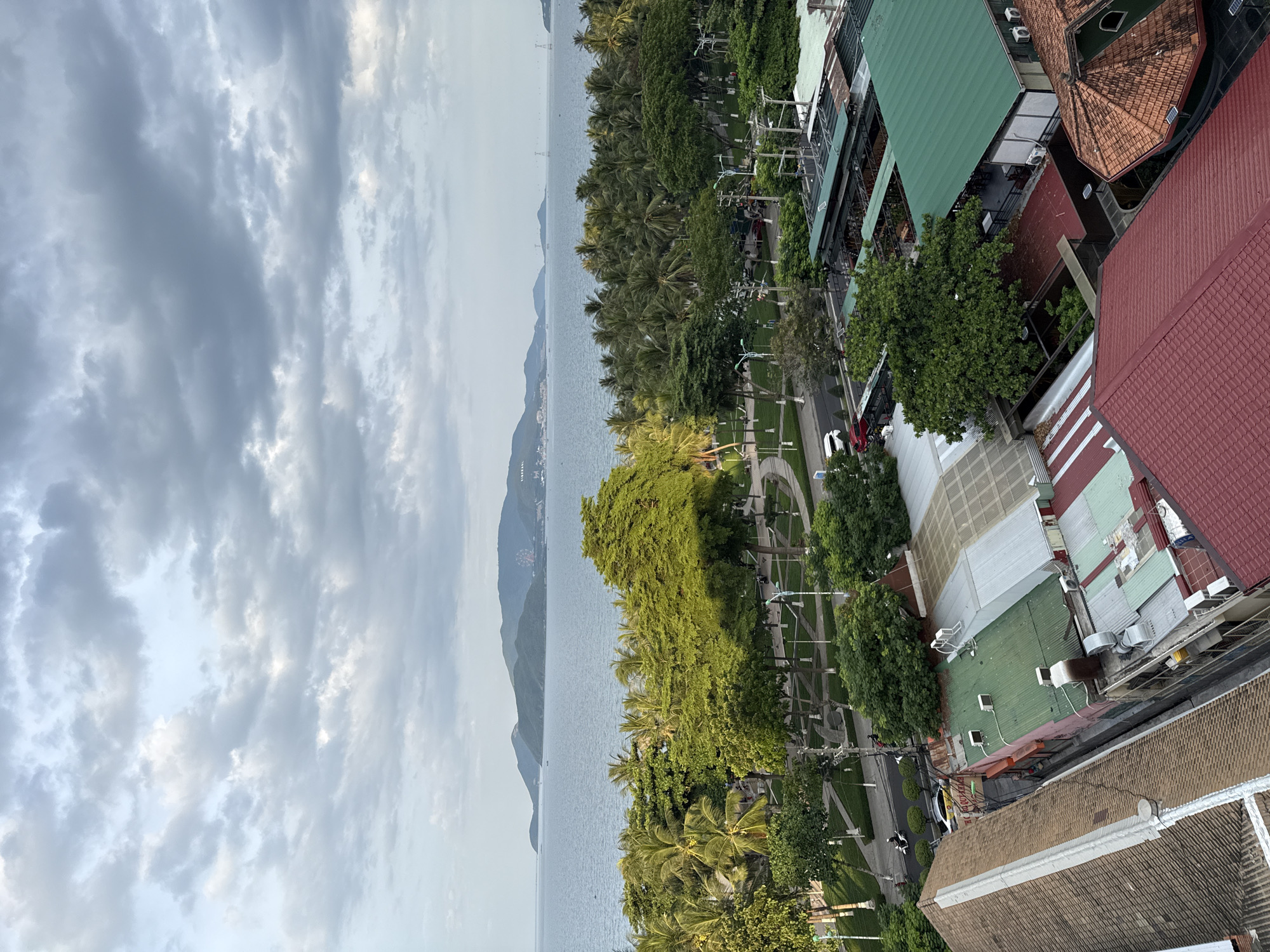 The image, taken from a high vantage point, captures a coastal city scene in Nha Trang, Vietnam, on a cloudy day. In the foreground, a cluster of buildings with red and green tiled roofs are visible, some with visible air conditioning units. Beyond the buildings, a tree-lined street is present, with several vehicles including cars and motorcycles, and pedestrians walking. Further back, a lush green park area with palm trees and other foliage lines the coast. The calm, blue sea stretches out to the horizon, meeting a hazy sky filled with gray clouds. In the distance, a chain of green, mountainous islands is visible, with a Ferris wheel and other structures on one of the larger islands. The overall impression is of a bustling coastal urban environment with a prominent natural landscape.