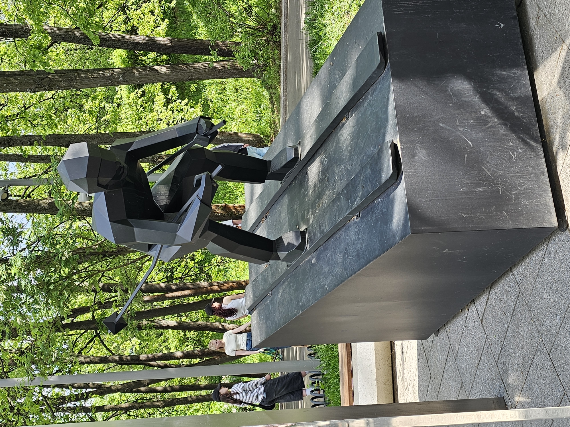 The image depicts a black, cubist-style sculpture of a skier in a seated position, located outdoors in what appears to be a park or garden setting. The sculpture is mounted on a dark gray, rectangular base. The skier figure is leaning forward with arms outstretched, holding ski poles. Behind the sculpture, there's a backdrop of trees with green foliage, suggesting a natural environment. Several people are visible in the background, appearing to be walking or sitting near the sculpture. There is a railing along the foreground, indicating a path or walkway. The sculpture may be located in the city of Moscow, Russia, as a relevant data-point.