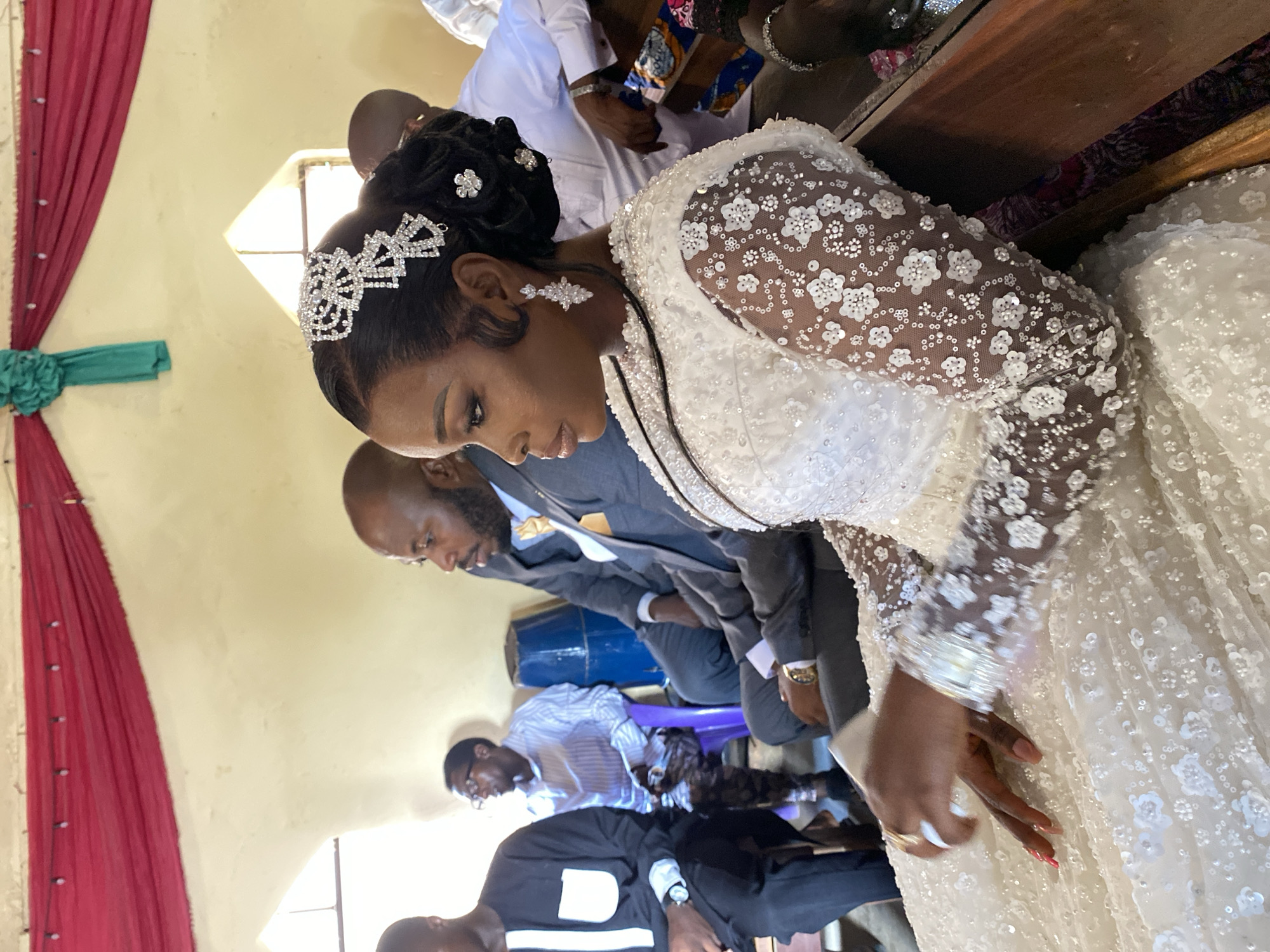 The scene depicts a wedding in Vom, Nigeria. The bride, adorned in a white gown with long, embroidered sleeves and a sparkling tiara, is the focal point. She stands near seated guests, including a man in a gray suit and gold tie. The setting appears to be indoors, possibly a church or hall, with red and green fabric decorations on the wall. Other attendees are also present.