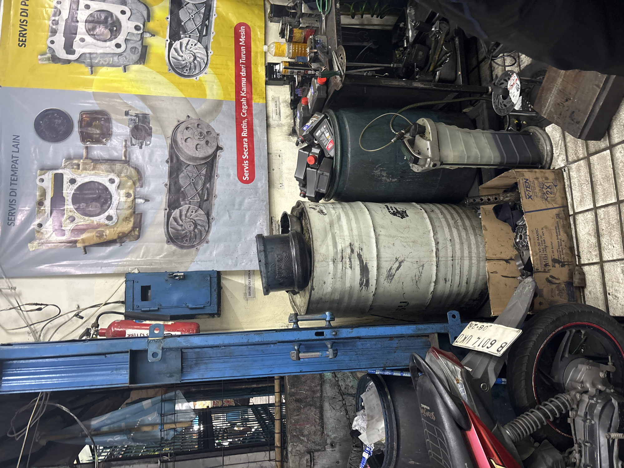 The image depicts the interior of a motorcycle repair shop, likely in North Jakarta, Indonesia. The scene is cluttered, with various tools, spare parts, and barrels of oil. In the foreground, a red and black motorcycle is partially visible, with its license plate reading "B 6014 UKD". Behind the motorcycle, there are two large, weathered drums, one white and one dark green, both appearing to be oil drums. A tall, grey oil drainage and collection unit stands next to the drums, with a hose connected.
On the wall behind the barrels, a large banner advertises motorcycle engine services, with images of engine parts and text in Indonesian. To the left of the banner, a blue electrical box is mounted on a blue metal post, with a red fire extinguisher hanging next to it. Various electrical wires are visible overhead. A workbench is set up to the right, laden with bottles of oil, batteries, and other repair equipment. A cardboard box containing more oil bottles is on the floor. The overall environment suggests a busy, working workshop, with evidence of ongoing maintenance and repair activities. The floor is tiled, and the lighting appears to be artificial, consistent with an indoor commercial space.