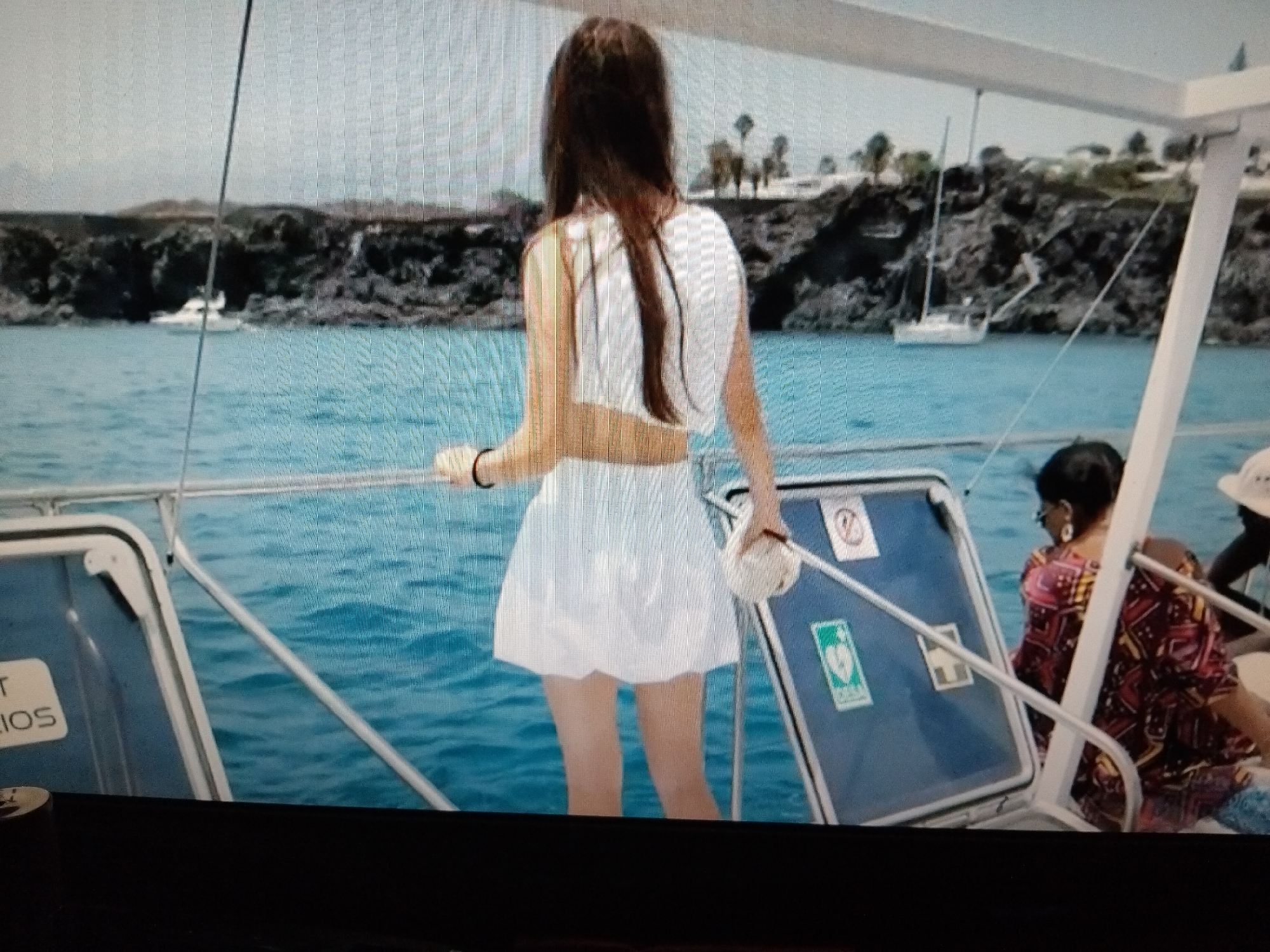 The image depicts a rear view of a woman standing on a boat, wearing a white crop top and a white skirt, facing a body of water. She has long dark hair and is holding a white object in her right hand. The boat appears to be on a turquoise sea, with a rocky coastline in the background, dotted with trees and some buildings. Another person, a woman wearing a colorful patterned dress and earrings, is seated on the boat behind the main subject, facing away from the camera. A third person, wearing a hat, is partially visible in the far right. There are also some safety signs on the boat. The sky is overcast and appears to be daytime. The scene is not in Bila Krynytsia, Ukraine, as it is a coastal scene with boats and a rocky landscape, not characteristic of that region.