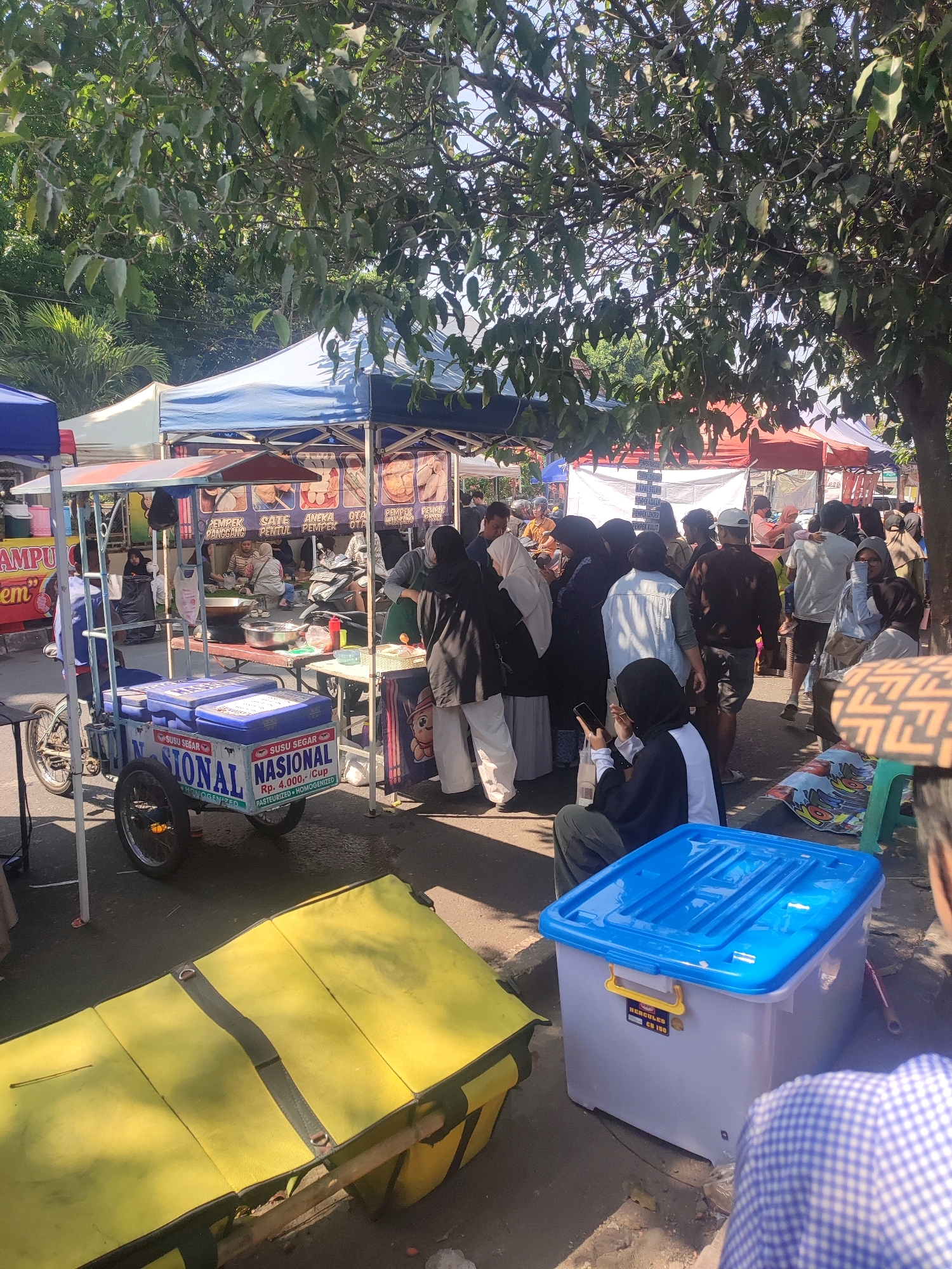 This image depicts a bustling street scene in Depok, Indonesia, likely a market or outdoor food stall area, under the shade of large trees. In the foreground, a yellow insulated carrier rests on the ground, and a blue plastic storage bin with a yellow handle is positioned to its right. Further back, a mobile ice cream cart labeled "SUSU SEGAR NASIONAL" with prices indicated in Rupiah is prominently displayed. Behind the cart, several food stalls are set up under tents, with awnings of various colors including blue, white, and red. Many people are present, some queuing at the stalls, others socializing, and a few are seated. A woman in a black headscarf and white shirt is seated in the lower right corner looking at her phone, while another person in a blue and white checkered shirt is partially visible. The overall atmosphere suggests a lively commercial setting on a sunny day.