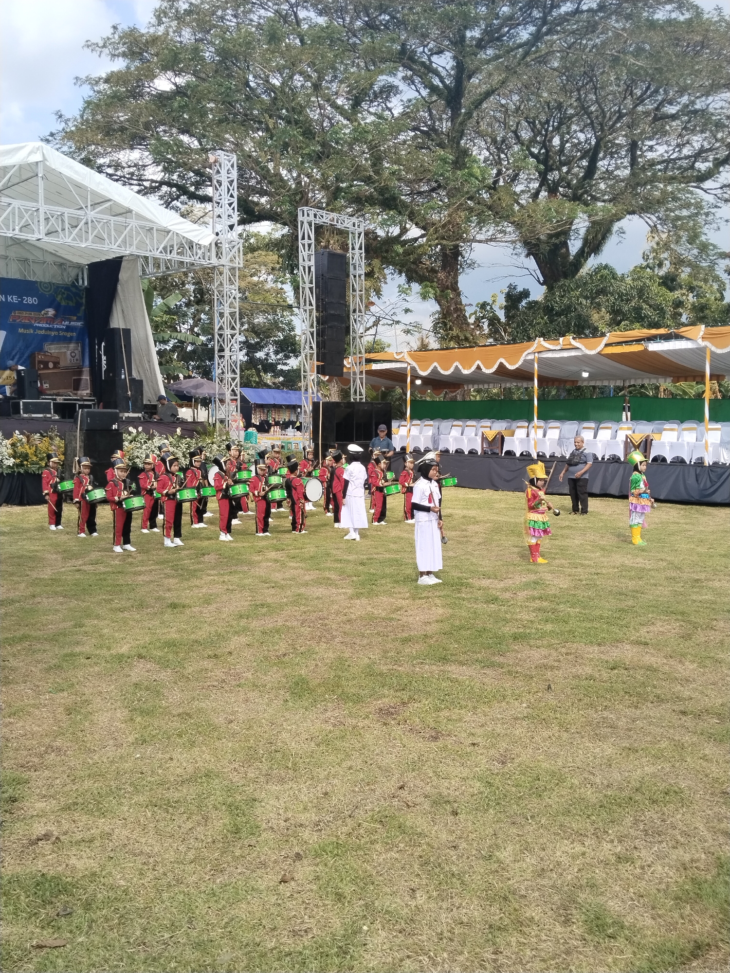 The image captures an outdoor scene in Sambirejo, Indonesia, featuring a marching band performance on a grassy field. The band consists of individuals dressed in red, green, and black uniforms with drums, and others in white uniforms with ceremonial swords. Two individuals in colorful costumes stand apart from the main group.
Behind the band, there is a stage with a white truss structure and speakers. To the right of the stage is a canopy structure with white chairs underneath. In the background, there is a large tree with dense foliage.
The scene appears to be a formal event or celebration. Several adults can be seen overseeing the performance. The weather is clear, with a partly cloudy sky.