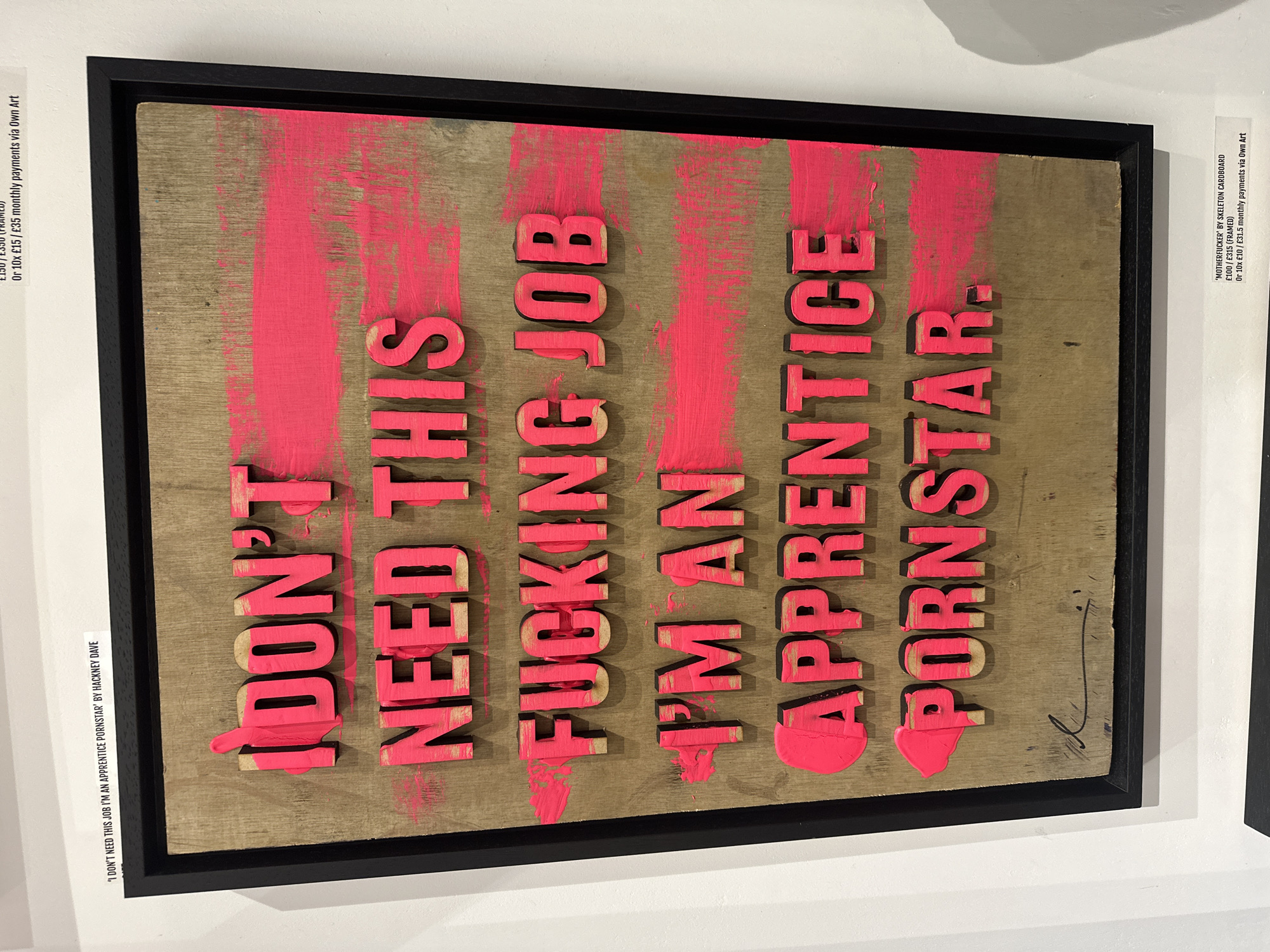 A framed piece of contemporary artwork is displayed on a white wall, likely in an art gallery or exhibition space in London. The artwork features three-dimensional block letters, painted in a bright, neon pink with visible drips and a slightly distressed finish, arranged against a raw, light brown wooden or cardboard panel. The text on the artwork reads: "I DON'T NEED THIS FUCKING JOB I'M AN APPRENTICE PORNSTAR." A black signature is visible on the bottom right of the panel.
The artwork is enclosed in a simple black frame. On the white wall, two labels provide contextual information. The partially visible label on the left identifies the piece as "'I DON'T NEED THIS JOB I'M AN APPRENTICE PORNSTAR' BY HACKNEY DAVE" and indicates pricing options such as "Or 10x £15 / £35 monthly payments via Own Art". A label on the right side details another artwork, "'MOTHERFUCKER' BY SKELETON CARDBOARD", with pricing "£100 / £315 (FRAMED)" and payment options "Or 10x £10 / £31.5 monthly payments via Own Art". The image is taken at a slight angle, showcasing the depth of the frame and the exhibition setting.