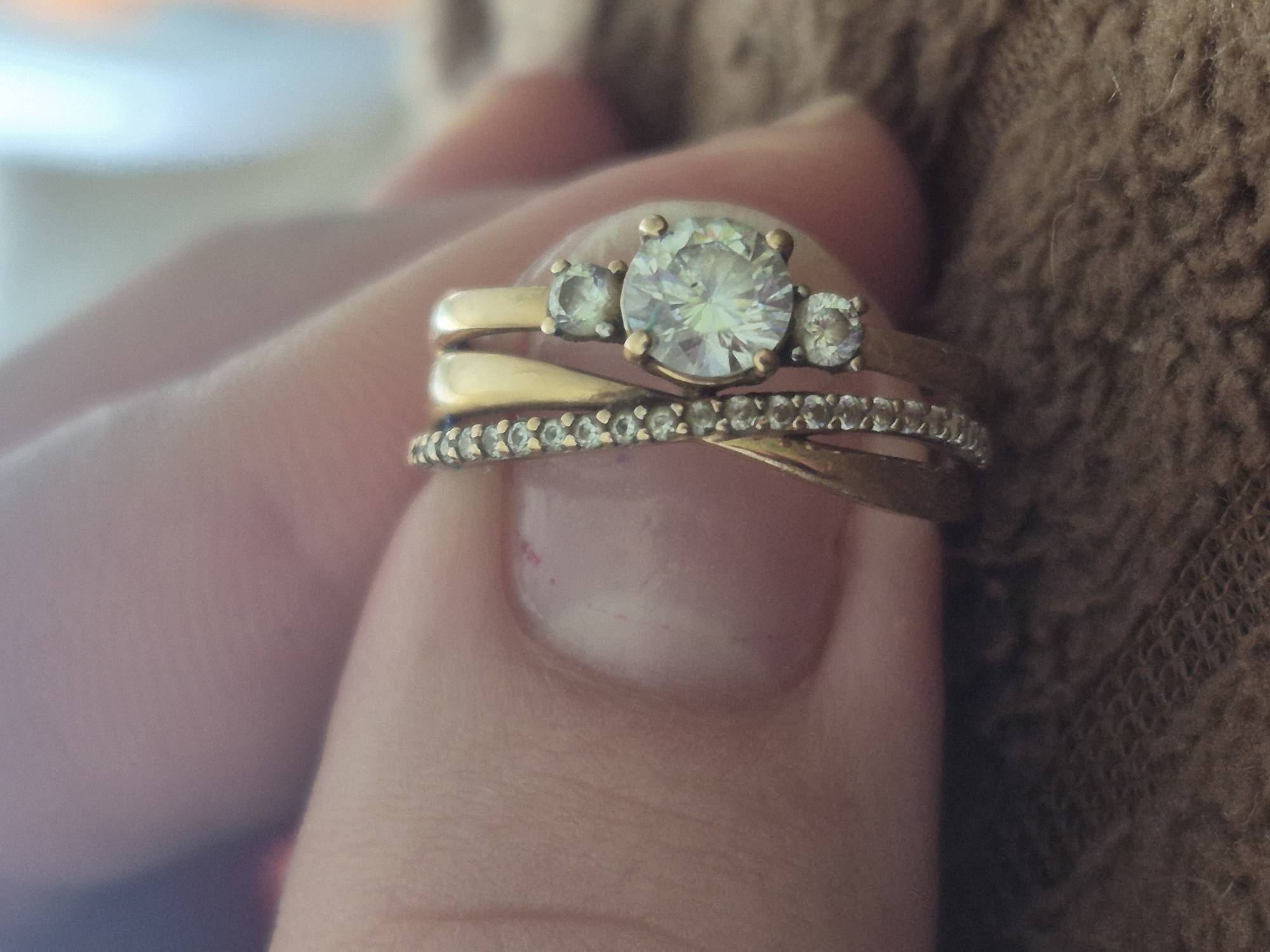 A hand, presumably female, holds three gold rings on the middle finger. The top ring features a large, central round stone flanked by two smaller stones. The middle ring is a plain gold band. The bottom ring is a gold band embedded with small stones. The background consists of a blurred tan surface, possibly clothing or fabric. There is no indication of Tobolsk, Russia or any location.