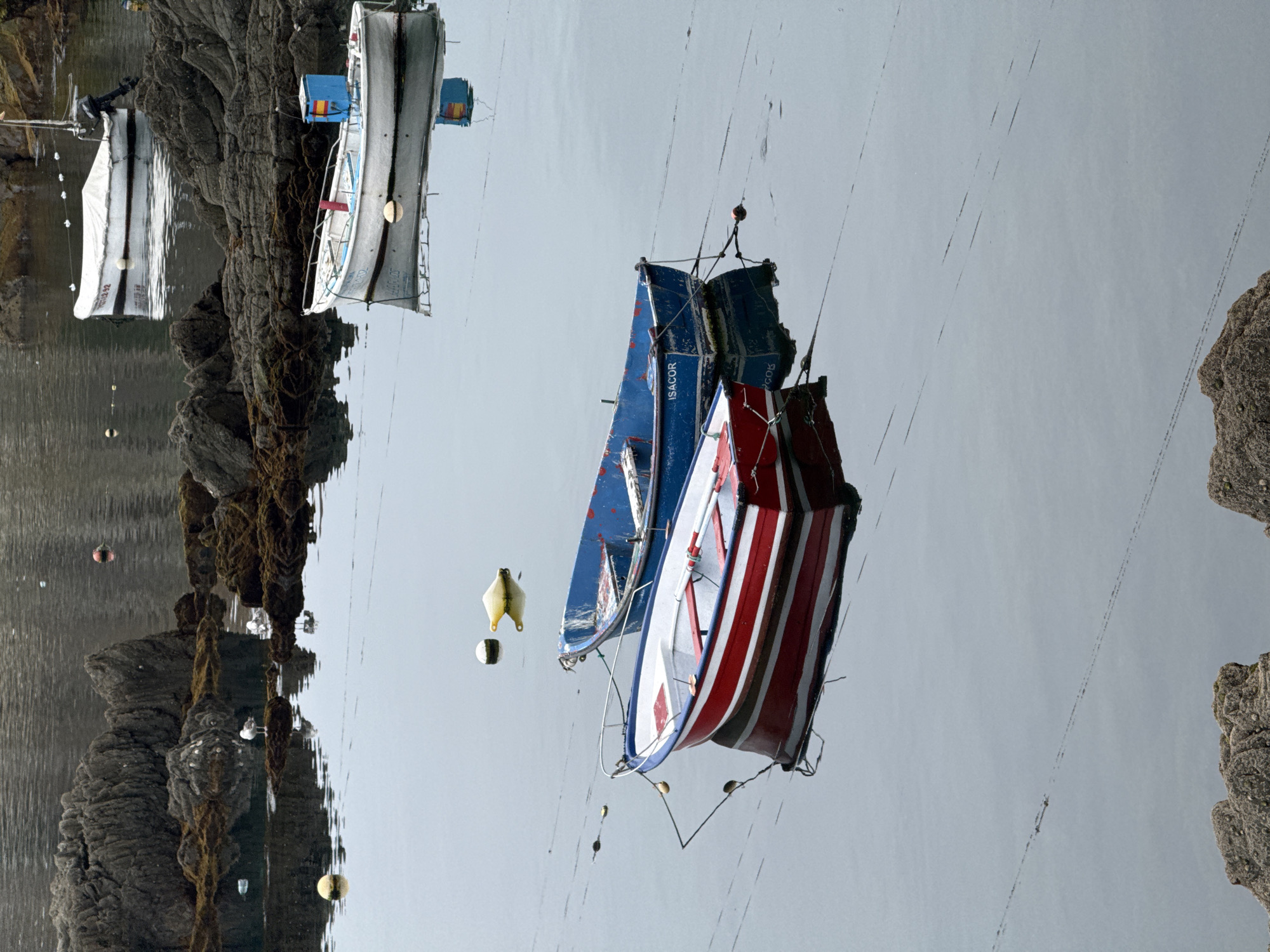 This media file shows a reflection of a harbor scene in Cudillero, Spain, as the image is upside down. The primary subjects are several small boats, some moored and others potentially on the water's surface.
In the foreground, a red and white striped boat is prominently featured, with a blue boat positioned behind it. Both boats appear to be of a style commonly used for fishing or leisure in coastal towns. There are also several buoys floating in the water, one yellow and one white with black markings.
Further back, on the left side of the image, a larger white boat is covered with a tarp and moored near a rocky shore. The rocky coastline is textured and dark, suggesting it's a natural formation typical of the region. The water appears calm, reflecting the sky and the objects above, creating a mirrored effect.
The scene is likely set during the daytime, with the lighting suggesting a bright, possibly overcast sky due to the soft reflections. The overall mood is tranquil and still. There is text visible on the blue boat which reads "ISACOR".