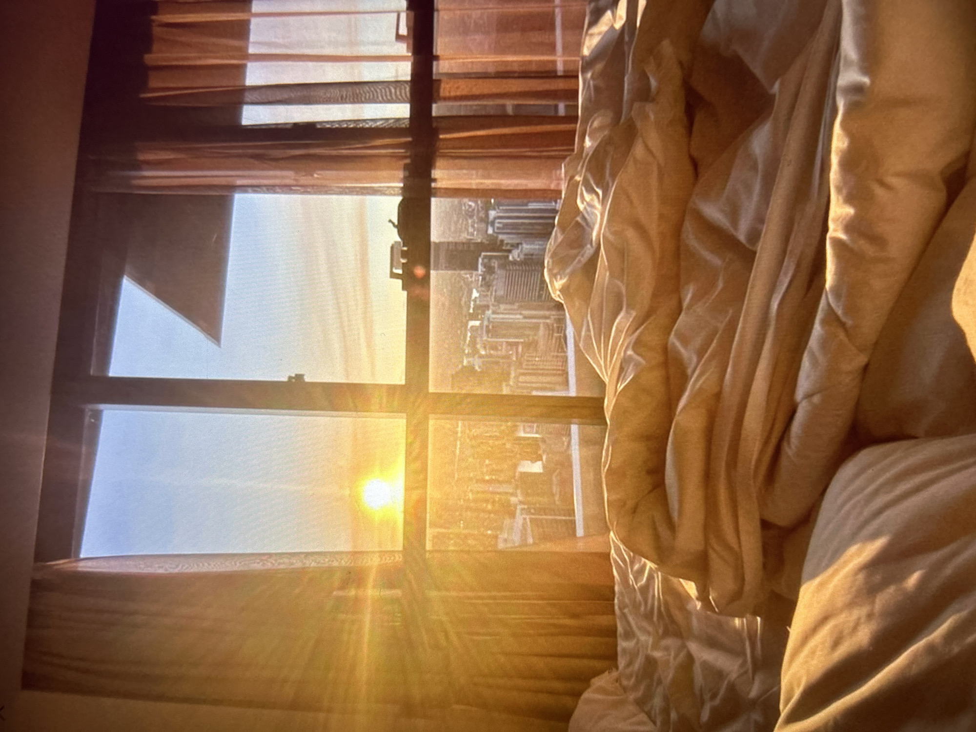 The image depicts a bedroom with a large window overlooking a city skyline at sunrise. The sun is prominently visible, casting a warm, golden light through the window and across the rumpled bedding. The room is furnished with a bed, partially covered by blankets and sheets, and sheer curtains hang on either side of the window. The city in the background shows a cluster of buildings, suggesting an urban environment. The overall impression is one of a serene morning scene. The prompt requests a description of the location in Knezha, Bulgaria; however, there are no visual cues in the image to confirm this specific location.