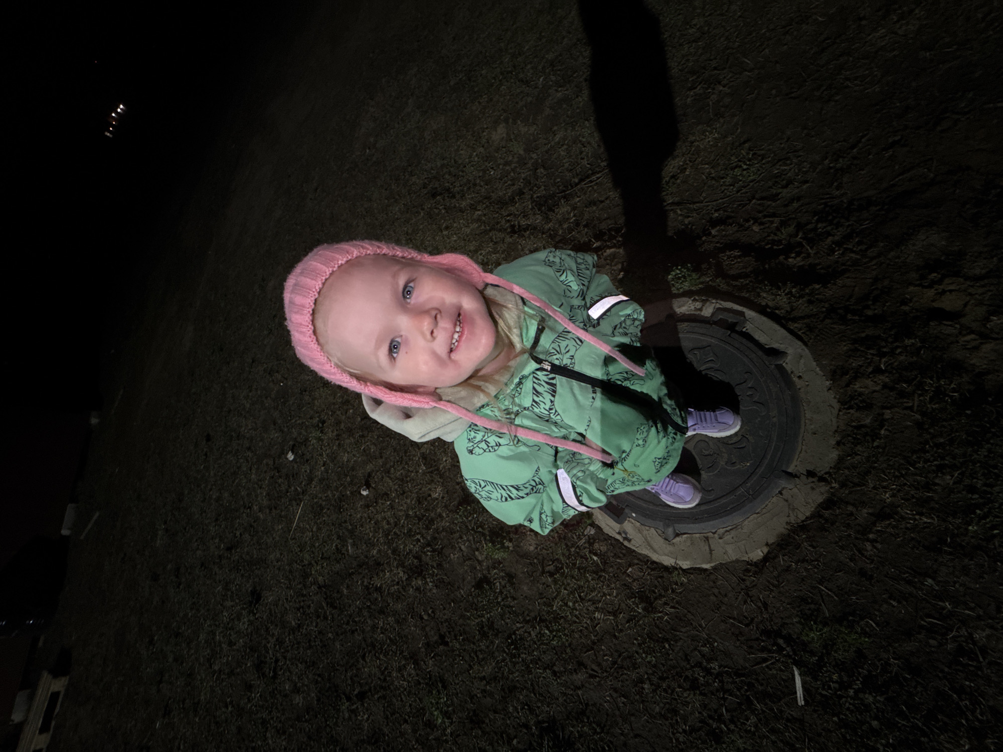 A young child stands on a manhole cover in Gornyi (Dvurechenskoe S/P), Russia. The child wears a pink knit hat and a green jacket with a tiger pattern. The child's shoes are purple. The surrounding environment is dark, possibly at night. Grass or dirt ground is visible. The lighting is dim, indicating the presence of a light source, with a shadow suggesting the direction of light.