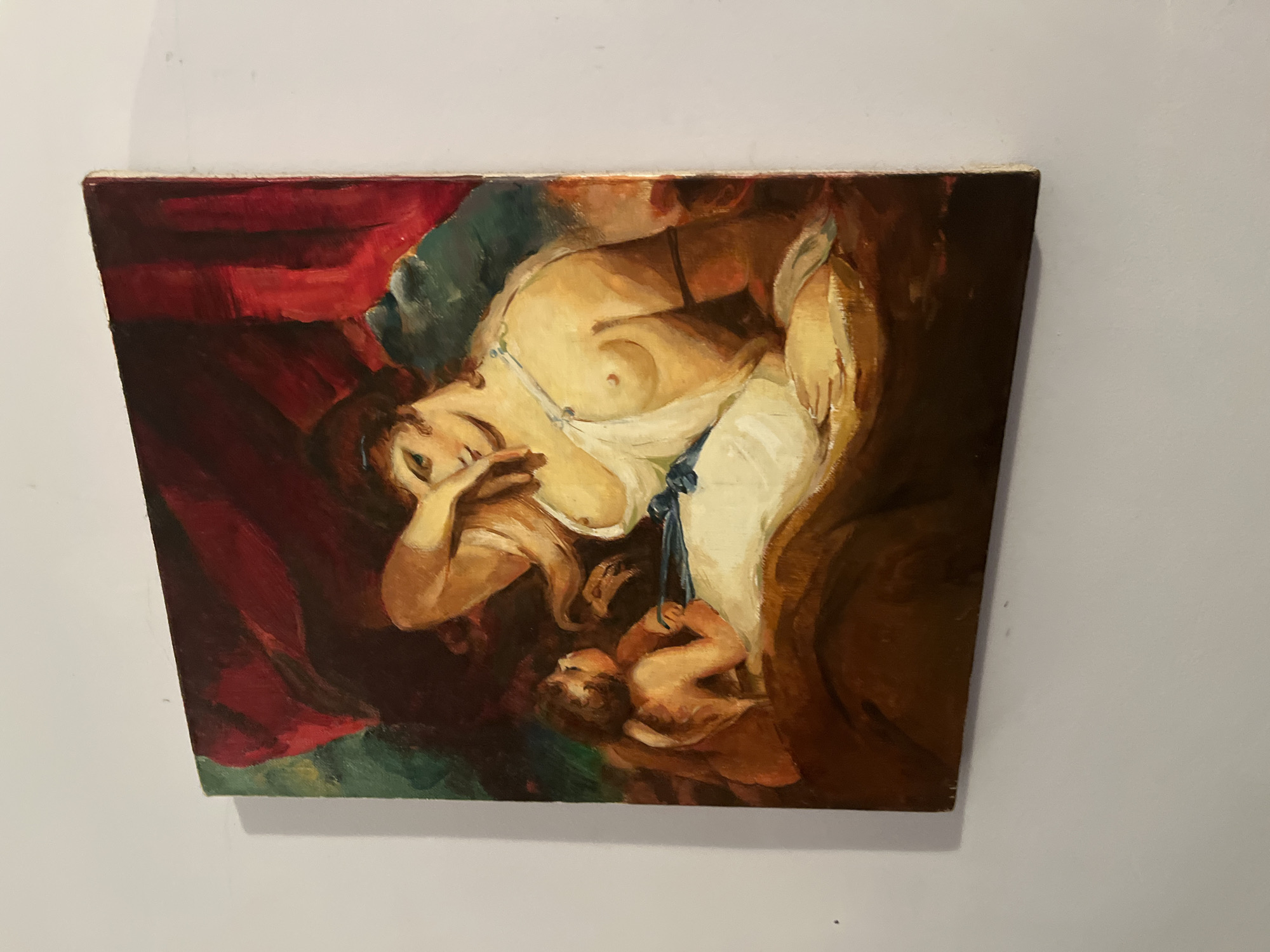 The image shows a painting hanging on a white wall. The painting depicts a woman lying down with a baby. The woman is fair-skinned and appears to be in a state of repose, with one arm raised to her face. She is partially unclothed, wearing a white garment with a blue tie. The baby is held close to her, also appearing fair-skinned. The background includes red and green drapery. The style of the painting is reminiscent of classical art. No specific location in Goce Delchev, Bulgaria is indicated, only that the painting is on a wall.