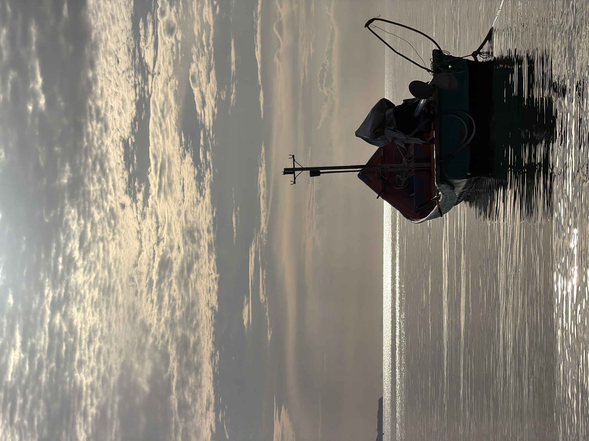 A single, small, unoccupied fishing boat is moored in calm, reflective waters, likely off the coast of Ko Samui District, Thailand. The boat, painted teal and black with red accents, carries a tarp-covered engine or equipment. A pole with two hooks stands prominently at the stern. The water's surface mirrors the overcast sky, which is filled with textured, layered clouds, illuminated by a diffused light source. In the distance, a faint landmass is visible on the horizon. The scene is serene and still, conveying a sense of early morning or late evening calm.