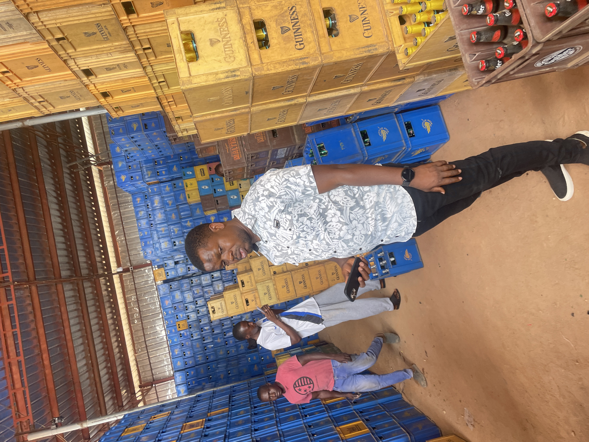 Three men are standing in what appears to be a large storage facility in Gura Top, Nigeria. The facility has a metal roof, and the floor is dirt. Stacks of blue and yellow plastic crates, many labeled "GUINNESS," fill much of the space. Some crates appear to contain beer bottles.
The man closest to the viewer is wearing a patterned gray and white shirt, black pants, and black and white sneakers. He is holding a black phone. The second man is wearing a white shirt with a blue stripe on the shoulder, gray pants, and dark sandals. The third man is wearing a pink shirt and blue jeans.