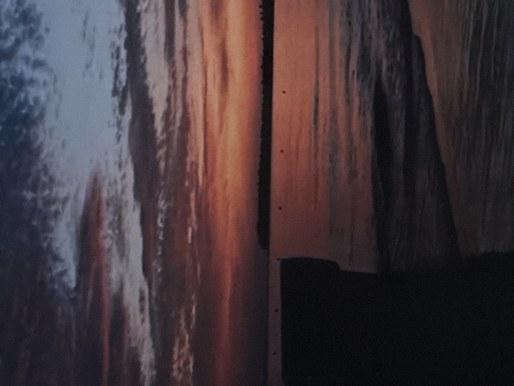 A nighttime photograph captures a coastal scene at dusk. In the foreground, a dark, blurry mass occupies the left side of the frame, obscuring details of its form, possibly a structure or a boat. The middle ground reveals a body of water with gentle waves lapping at an unseen shore. The water reflects the muted colors of the sky. In the distance, a thin, dark line suggests a distant shoreline or a low-lying landmass. The sky above is a canvas of twilight, with streaks of deep purple, fiery orange, and pale blue clouds. Two small, indistinct red buoys are visible in the water, near the distant shoreline. The overall impression is one of tranquility and the fading light of day.