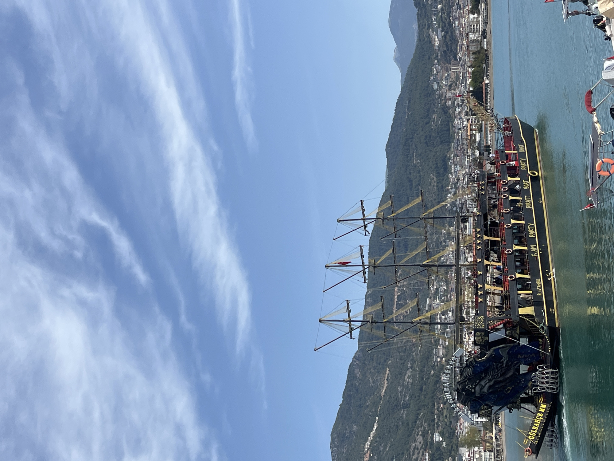 The image captures a docked pirate-themed "party boat" called "Gülbahçe MM" in Alanya, Turkey. The vessel, predominantly black with gold trim, features multiple masts, likely constructed for decorative purposes rather than sail functionality. The vessel is labeled with "Piratt Boat Party," suggesting its function for tourist entertainment. The boat is docked in a harbor; the sea is calm. Behind the boat, the landscape shows a densely populated area ascending a hillside, indicative of the city's terrain. Mountains rise further in the background. The sky is clear with wispy clouds. The scene suggests a tourist destination in Alanya, with the pirate ship likely offering excursions along the coast.