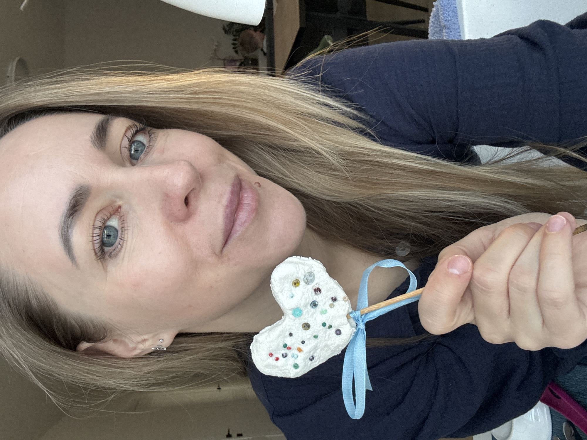 A light-skinned woman with long, blonde hair and striking blue eyes looks directly towards the viewer with a subtle, pleasant smile. She is positioned diagonally across the frame, wearing a dark blue long-sleeved top. In her right hand, held at chest level, she presents a handmade, heart-shaped object on a light brown wooden stick. The heart is white with a textured surface, decorated with numerous small, colorful beads or sequins in various shades including blue, green, yellow, red, purple, and silver. A light blue ribbon is neatly tied around the wooden stick below the heart, forming a bow.
The background is softly blurred, suggesting an indoor setting such as a home or craft area. Visible hints in the background include a white roll of paper or fabric, a green plant, and a light blue textured item. Natural, soft lighting illuminates the scene, indicating it is daytime. The woman's expression is calm and engaging. Notable details on her include small ear piercings and a small mole above her upper lip. The activity appears to be the presentation of a handcrafted decorative item. No visible text is present in the image.
Location context: Chaikovsky, Russia.