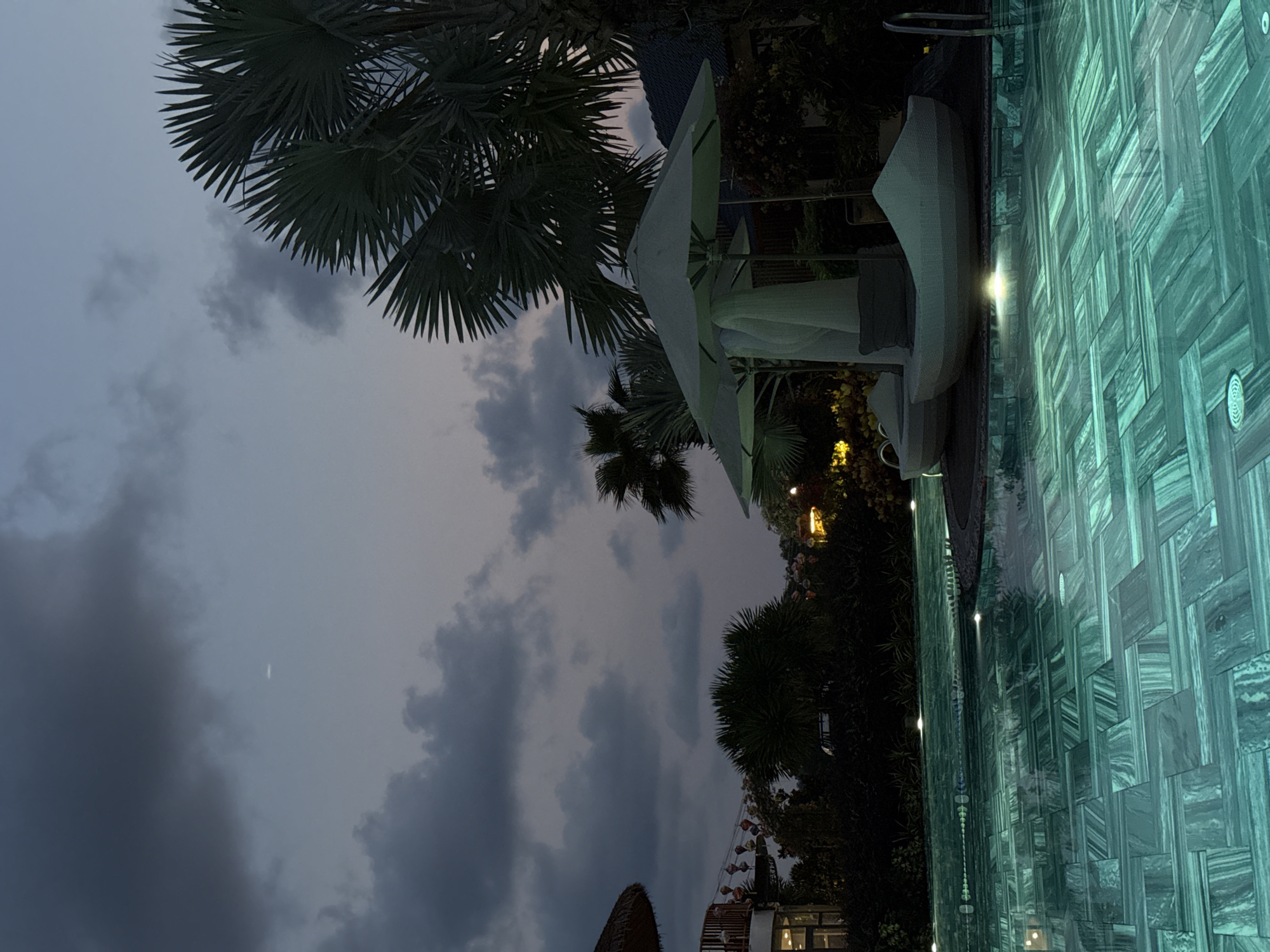 The image captures an outdoor evening or nighttime scene at a resort in Phú Quốc, Vietnam, featuring a swimming pool, lounge furniture, and tropical vegetation under a cloudy sky.
In the foreground, a vibrant, illuminated swimming pool dominates the right side of the frame. The pool water glows with a turquoise or blue-green hue, and its bottom is lined with distinctive rectangular tiles in various shades of teal and gray, arranged in a horizontal, stacked pattern. Several bright lights are visible beneath the water's surface, creating reflections.
Along the edge of the pool, several modern, white outdoor lounge chairs or daybeds are arranged, some with canopy-like structures providing shade or privacy. A large, white patio umbrella is also prominently featured, open and casting a light shadow.
Behind the pool and furniture, lush, dark green tropical foliage is visible, with several large palm trees silhouetted against the sky. Their fan-shaped fronds are distinct against the lighter parts of the sky. Other dense vegetation and some twinkling warm lights suggest decorative lighting or nearby resort structures.
The sky is dark and cloudy, indicating dusk or nightfall. Patches of lighter gray sky are visible between the dark clouds, and a very faint, slender crescent shape, possibly the moon, can be seen in one of these lighter areas.
No people are visible, and no specific activity is taking place, contributing to a serene and tranquil atmosphere. No visible text is present in the image.