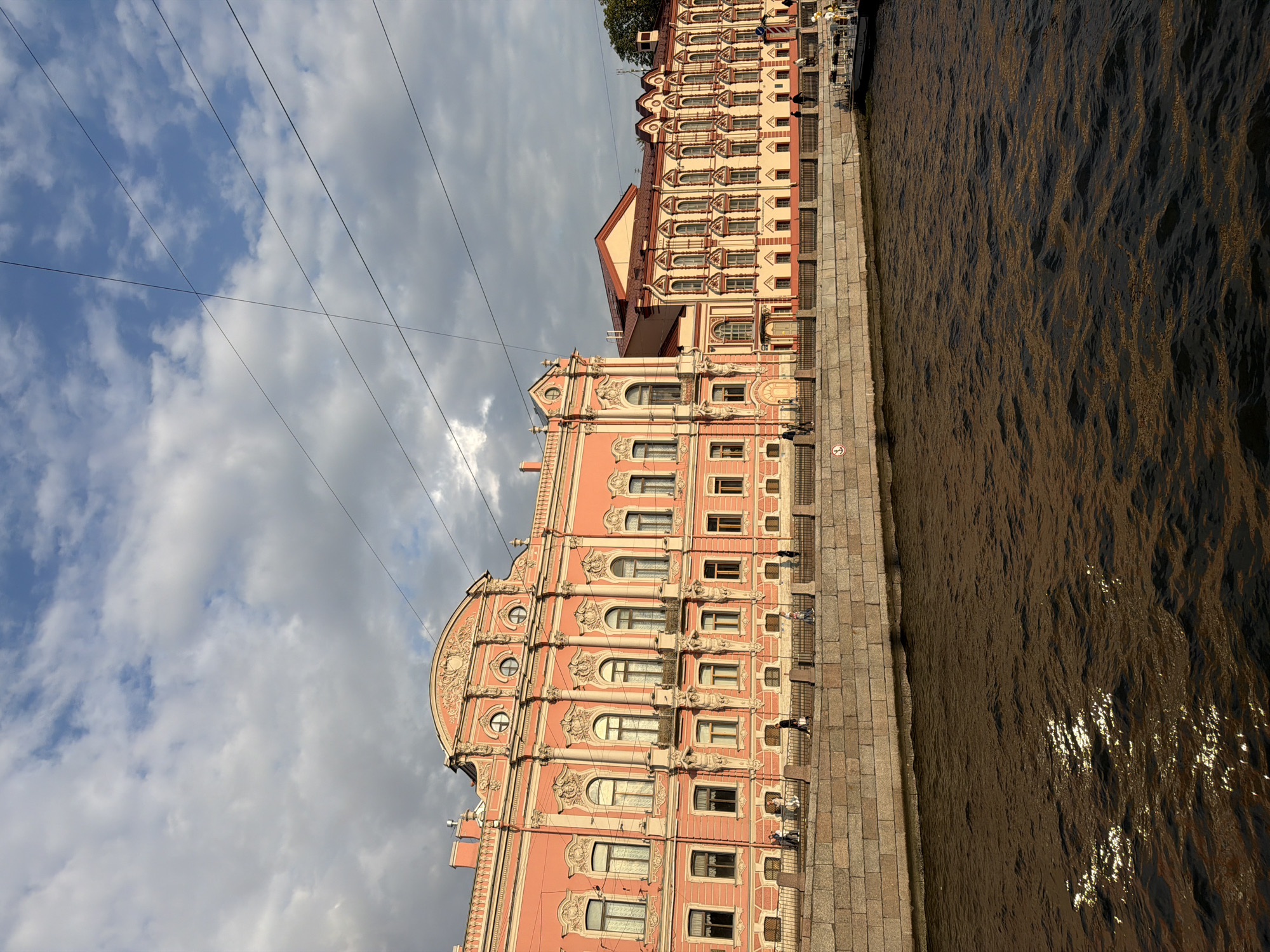 The image shows a wide view of the Moika River in Saint Petersburg, Russia, with two ornate, pink and cream-colored buildings lining the embankment. The architecture is elaborate, featuring arched windows, decorative columns, and intricate moldings. Several people are visible walking along the stone embankment, which is separated from the river by a metal railing. The river itself is dark brown with ripples and reflections of sunlight. In the distance, a white boat is docked along the embankment. The sky is a mix of blue and grey clouds, with visible power lines crisscrossing the upper portion of the frame.