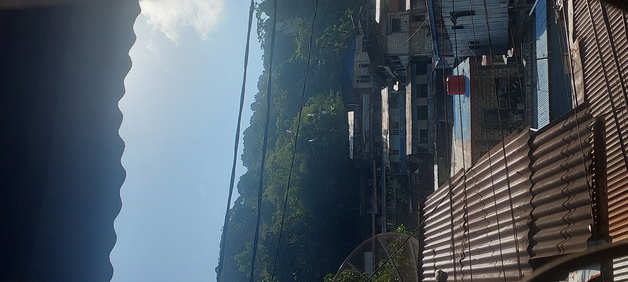The image captures a view of a densely populated hillside settlement in Jayapura, Indonesia, on a bright, clear day. The foreground is dominated by the corrugated metal roofs of several houses, some appearing aged and weathered. A large satellite dish is partially visible on the left.
Further into the scene, the settlement climbs up a lush, green, and densely forested hillside. Numerous buildings, constructed from a mix of brick, concrete, and metal sheeting, are stacked closely together. Many of these structures have corrugated metal or brightly colored (blue and red) roofs. Several utility wires are strung across the scene, partially obscuring the buildings and the hillside. A bright red water tank sits on top of one of the buildings.
The sky is a clear, bright blue with a few wispy white clouds visible, suggesting fair weather. The sunlight casts distinct shadows, highlighting the texture of the roofs and the uneven terrain. There are no people clearly visible in the image, nor any specific actions or interactions taking place. The overall impression is a snapshot of a vibrant, working-class community nestled against a natural, verdant backdrop.