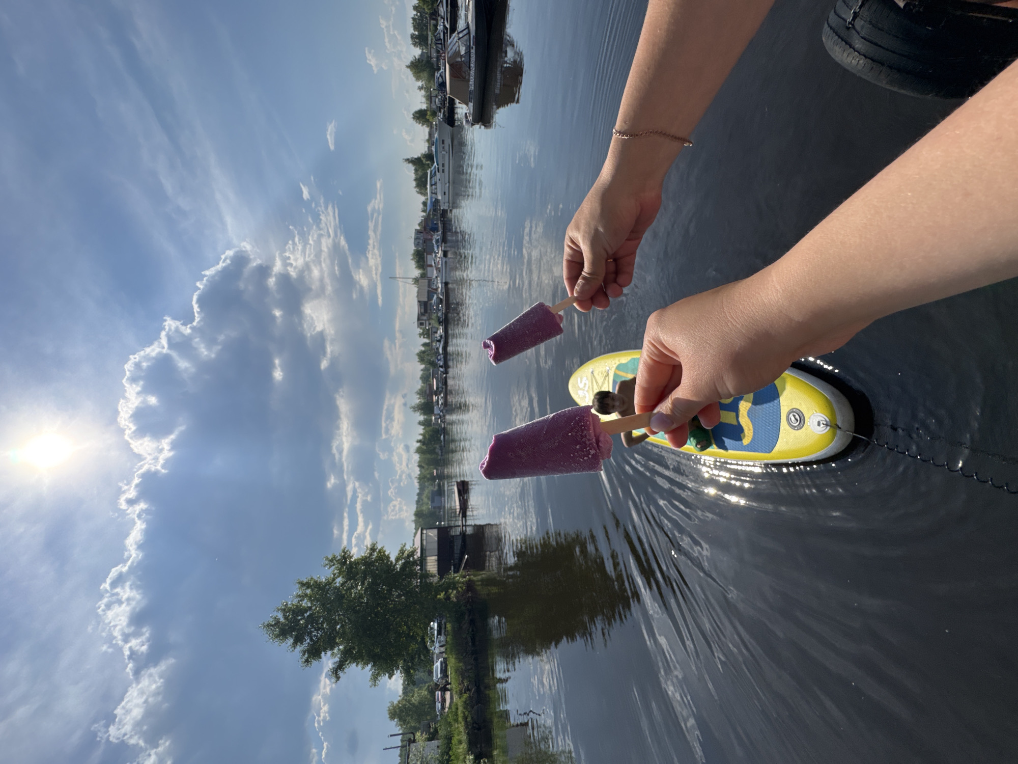 In Chaikovsky, Russia, on a bright, sunny day with scattered clouds, two hands hold purple popsicles over a calm, dark body of water, likely a reservoir or river. The popsicles are positioned in the foreground, with one held closer to the camera. In the background, a young person is seated on a yellow and blue paddleboard, which is moving through the water, creating ripples. The surrounding environment includes a treeline with a single large tree, several buildings that appear to be boat houses or small structures along the shore, and numerous boats docked in the distance. The sky is a vibrant blue with sunlight shining through gaps in the clouds.
