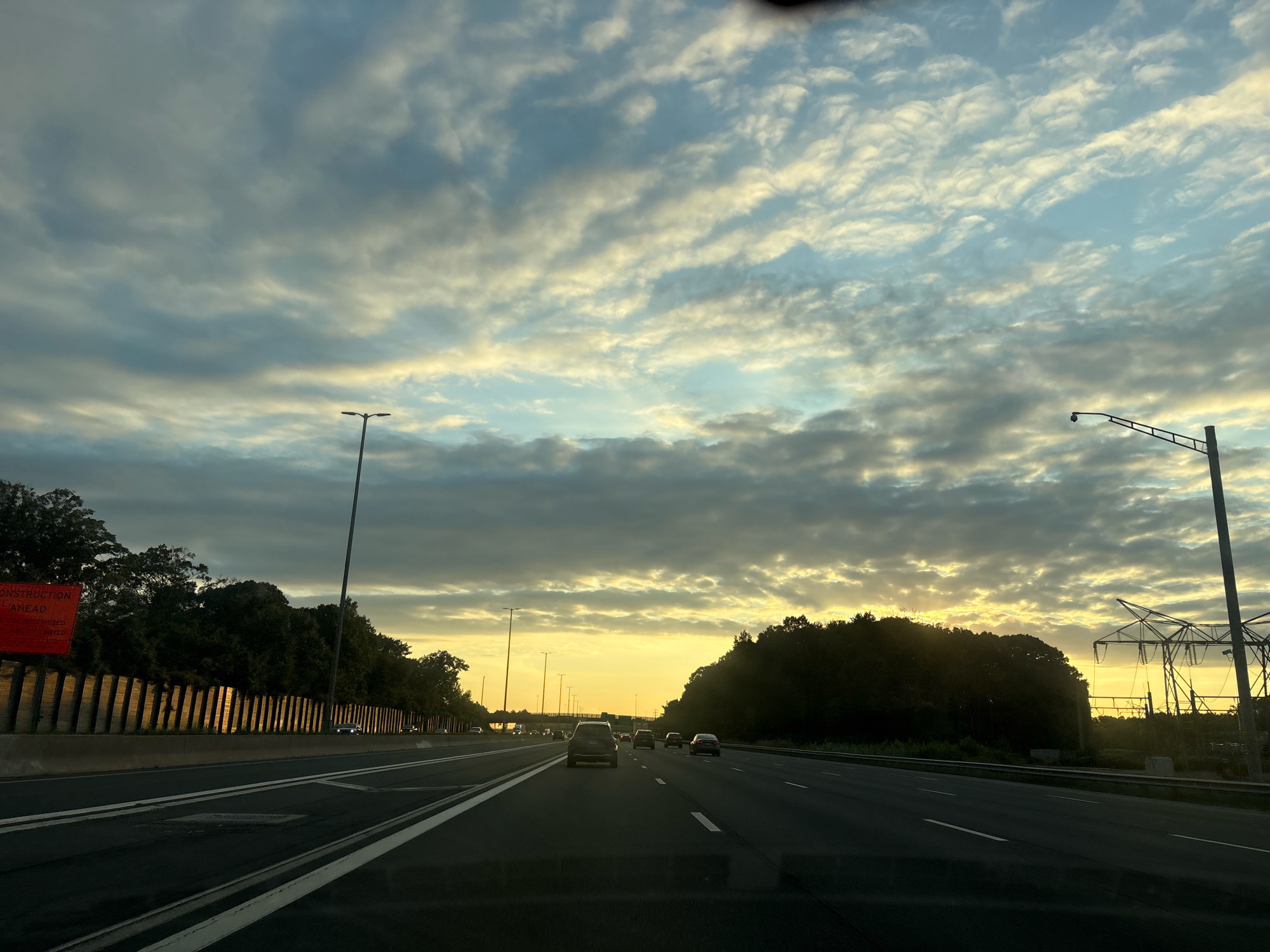 The image depicts a highway scene in East Hartford, Connecticut, United States of America, during a sunset. The sky is filled with dramatic clouds, transitioning from grey to yellow and blue as the sun dips below the horizon. A highway with multiple lanes is visible in the foreground, with cars driving in both directions. On the left side of the highway, there are trees and a construction sign. On the right side, there are more trees and a power substation with tall electrical towers. The overall atmosphere is one of a late evening commute as day turns into night.