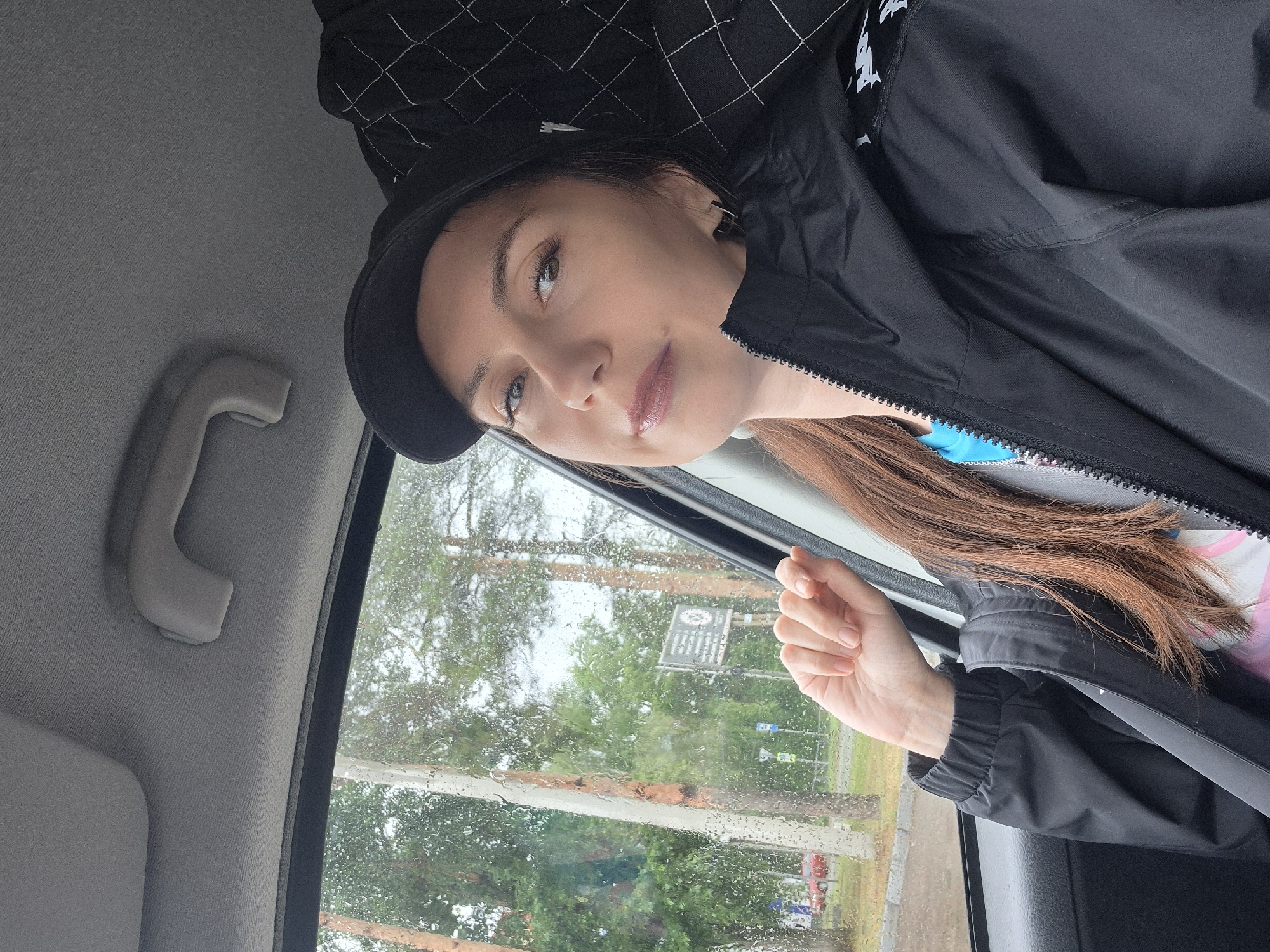 A medium-shot selfie shows a woman in a car, looking toward the camera. She is wearing a black baseball cap and a black jacket over a bright blue shirt. Her right hand is resting on the car door, and her left arm is bent, with her hand near her face. Raindrops are visible on the car window, and trees and a sign are blurred in the background outside the car. The interior of the car, including a gray headliner and a grab handle, is visible in the upper portion of the image.