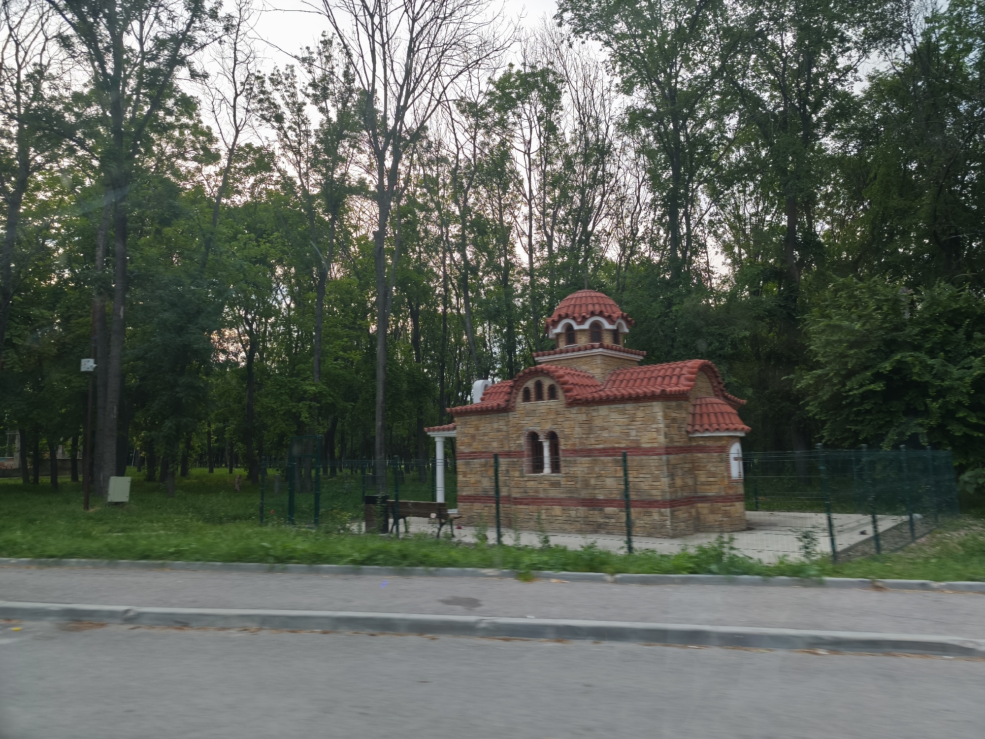 This image presents a small, ornate building with a distinctive red tiled roof, resembling a miniature church or chapel, nestled within a lush green park-like environment. The building is constructed with light-toned stone blocks and features reddish-brown brick accents, multiple arched windows or openings, and a small central dome. A dark green metal fence encircles the immediate perimeter of the structure, and a wooden bench is visible within this fenced area to the left of the building.
The background is dominated by a dense array of trees, some boasting full green canopies while others exhibit bare branches, possibly indicating early spring, late autumn, or specific tree types. The ground beneath the trees and around the building is covered in vibrant green grass. In the foreground, a paved road with a gray concrete curb runs horizontally across the frame.
The lighting is soft and diffused, suggesting either an overcast day or the gentle light of late afternoon or early evening. The scene is devoid of any people or active events, conveying a sense of peace and stillness. No visible text is present in the image. The architectural style of the small building, particularly its roof and stone masonry, hints at potential Southern European or Balkan influences.
