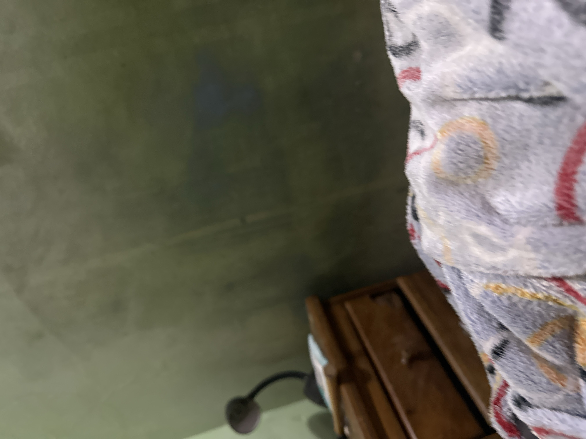 The image presents a blurry, low-angle interior view, dominated by a muted color palette. In the immediate foreground, a soft, textured textile object, possibly a blanket or clothing, is visible. It features a predominantly white or light gray base with abstract patterns including red curves, black geometric shapes, and yellow/orange circles.
To the left of the frame, partially obscured by the foreground textile, stands a wooden nightstand or small dresser with at least two visible drawers, each equipped with a small wooden knob. On the surface of this furniture, a dark-colored, gooseneck-style desk lamp is present, positioned next to a small, light-colored rectangular object, possibly a stack of papers or a book.
The background consists of a plain wall, painted in a desaturated olive-green or grayish-green color, exhibiting slight variations in tone and texture. No distinct features or decorations are discernible on the wall. The overall scene is softly lit, suggesting an indoor environment, likely a bedroom. No individuals or actions are visible within the frame. The specific geographical location, such as Juan Griego, Venezuela, is not discernible from the visual information provided in the image.