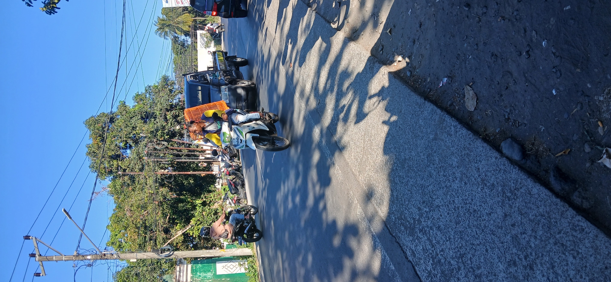 The image depicts a street scene in Kediri, Indonesia, under a clear blue sky. In the foreground, a man is riding a white scooter, carrying a large, orange, cage-like structure on the back. He is wearing a yellow and blue jacket and blue jeans. To his left, another person on a black scooter is riding away from the viewer, wearing a beige hoodie and jeans. Behind the white scooter, a dark-colored pickup truck is visible. Several other motorcycles are parked in the background, lining the side of the road. Trees with green foliage line the street, and power lines crisscross the sky. The road surface is asphalt, with some areas showing wear. Shadows of trees and vehicles are cast across the road, indicating it is daytime. The environment suggests a typical urban or semi-urban setting in Indonesia.