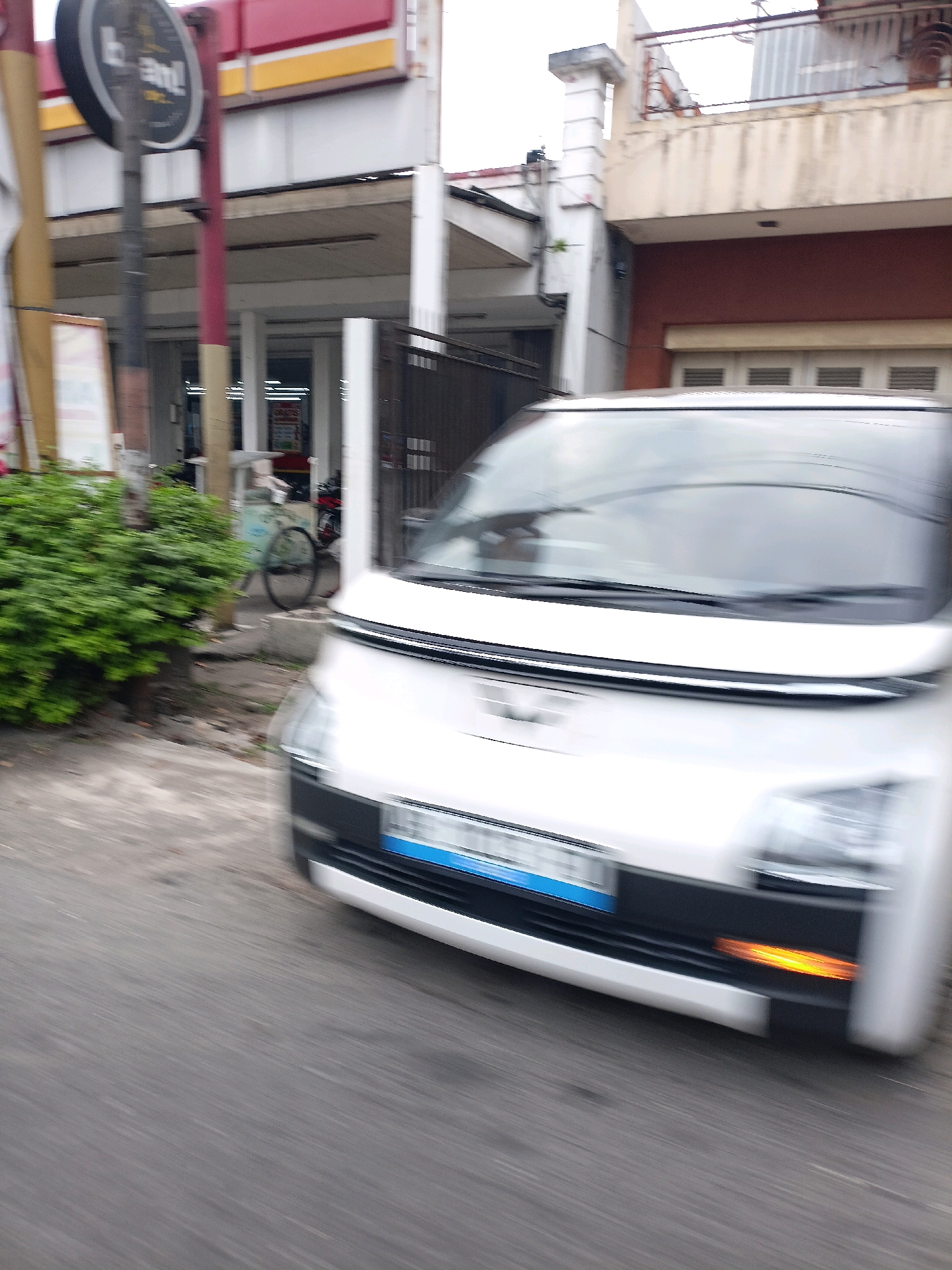 A white, four-wheeled vehicle is captured in motion on a paved road, likely in Magelang, Indonesia. The image is blurred due to movement, with the front of the vehicle prominently featured. The vehicle has a black grille and a blue license plate with white lettering. An orange turn signal is visible on the right side of the front bumper. To the left of the vehicle, a densely foliated green bush is present. In the background, a commercial building with a red and yellow awning is visible, along with a sign that appears to be circular and black with white text. A bicycle with a basket is parked near the building. Further back, there is a multi-story building with a balcony and tinted windows. The environment suggests an urban or commercial street setting.