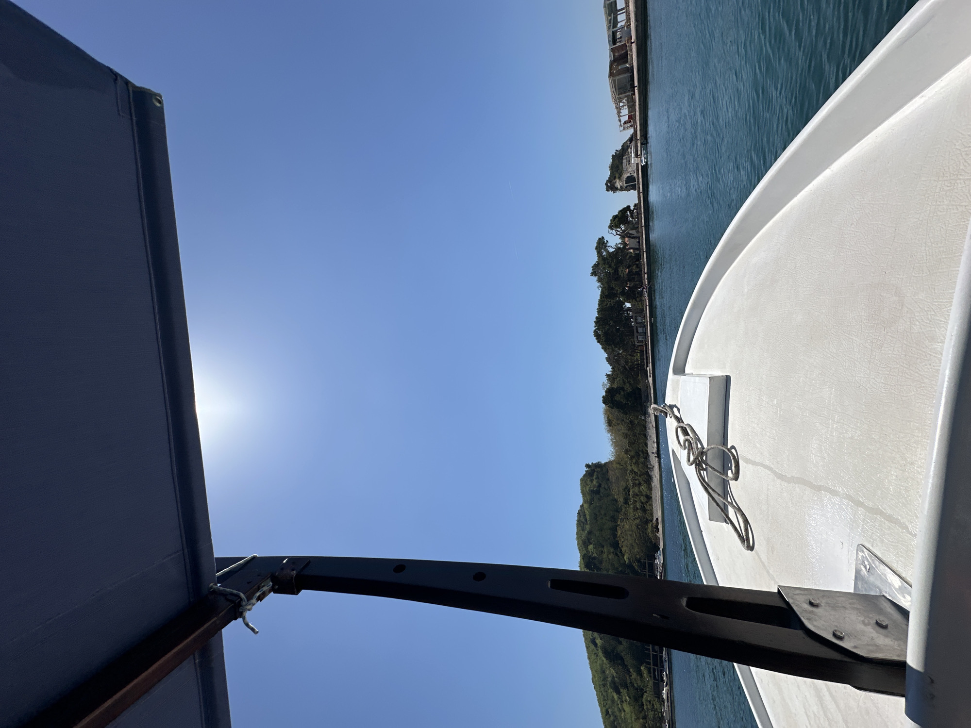 The photograph is taken from the perspective of someone on a boat. The sky is a clear, light blue with the sun visible behind an awning on the left. The water is calm and a deeper blue. A stretch of land is visible in the distance with trees and buildings. The side of the boat is white, and a black awning support is visible. The location is likely near Budva, Montenegro, given the coastal landscape and architecture.