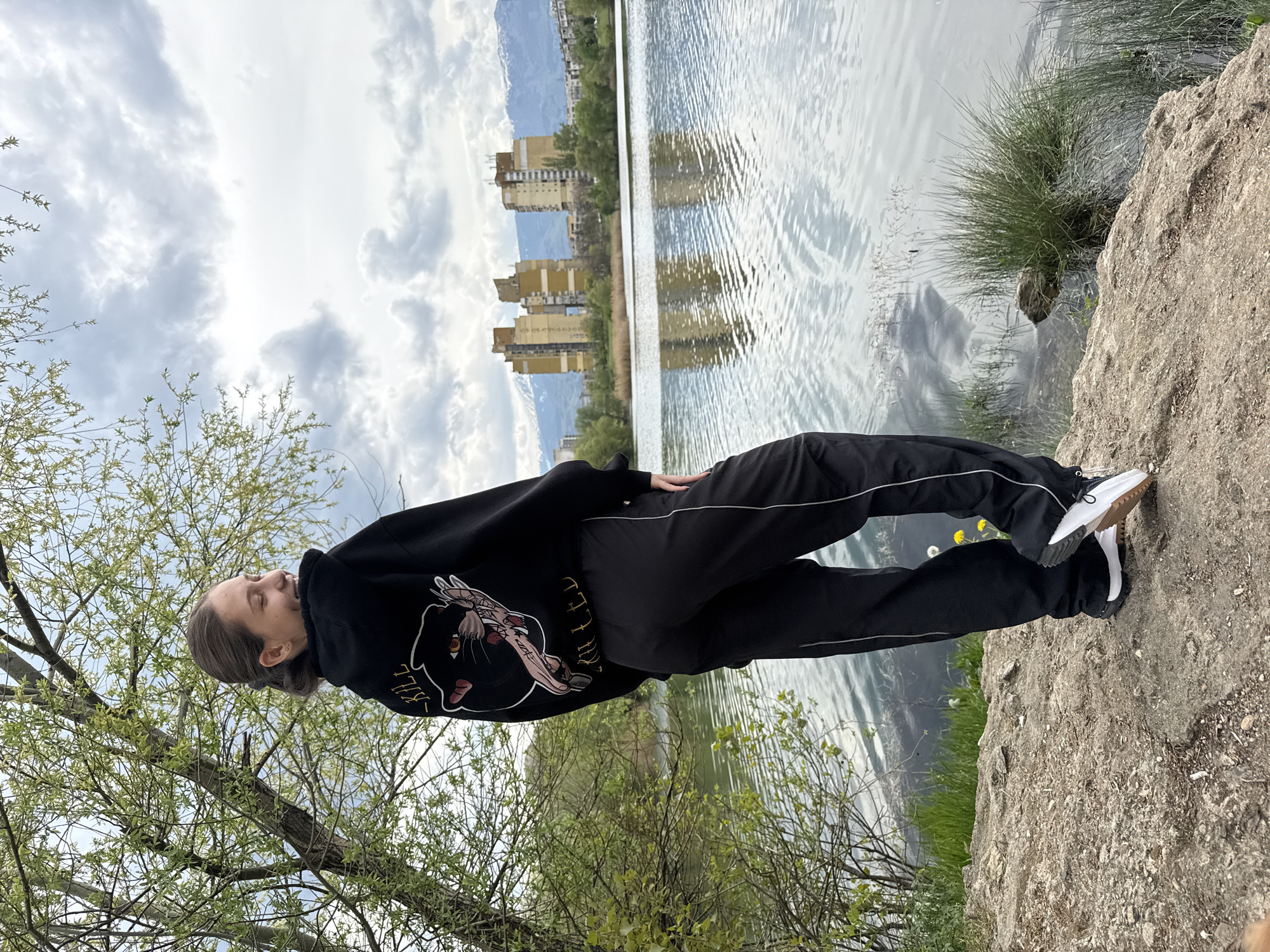 The image presents an outdoor scene of a person standing on a rocky outcrop beside a body of water under an overcast sky in Sofia, Bulgaria. The subject, a young adult with medium-length hair, is seen in profile, looking upwards with their eyes closed or nearly closed, conveying a sense of peaceful contemplation. They are wearing a black hooded sweatshirt adorned with a distinctive graphic of a winged, feline-like creature, with the word "KILL" prominently visible near the design. Black track pants with white stripes down the sides and white sneakers complete their attire. One hand is tucked into a pants pocket, while the other rests casually by their side.
The setting features a calm body of water, likely a lake or large pond, reflecting the muted light of the sky. Across the water, several multi-story buildings, possibly residential or industrial, stand in a line, characterized by their uniform, somewhat aged appearance. Beyond these structures, distant mountains rise, their silhouettes softly defined against the cloudy backdrop. The immediate foreground includes the rough, light-colored rocky ground where the person stands, along with patches of vibrant green grass and the budding branches of a tree on the left. The sky is dominated by a blanket of soft, grey and white clouds, suggesting a diffused, even light without direct sunlight. The overall atmosphere is tranquil, blending natural elements with an urban edge.