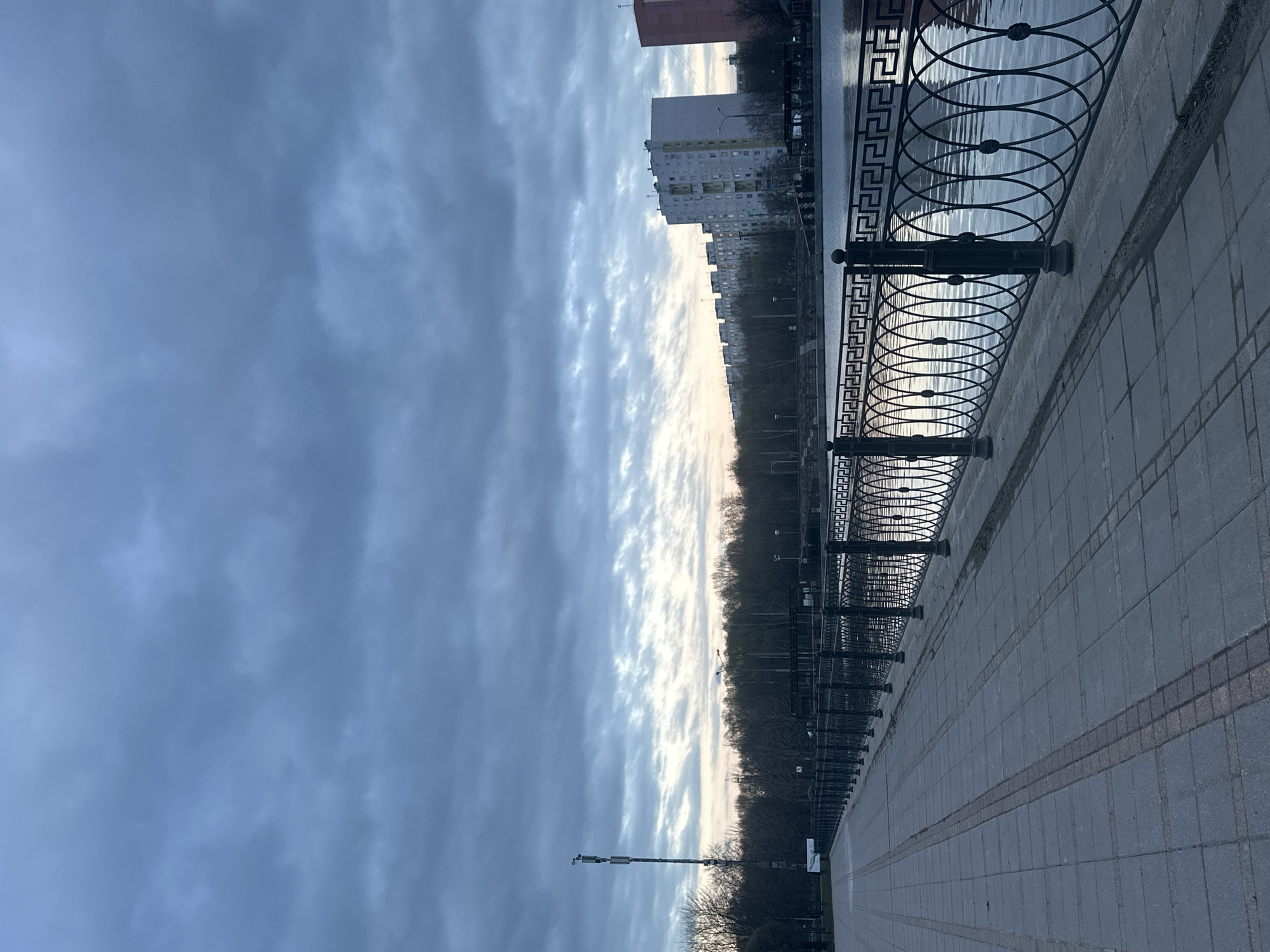 The image shows a riverside walkway in Moscow, Russia, under a cloudy sky. A decorative black iron fence runs along the edge of the walkway, separating it from the water. Behind the fence, the water reflects the sky and some distant buildings.
On the other side of the water, there are buildings and trees. A tall building is visible on the left side. The walkway is paved with grey stone. The lighting suggests either early morning or late evening. No people are visible in the immediate foreground.