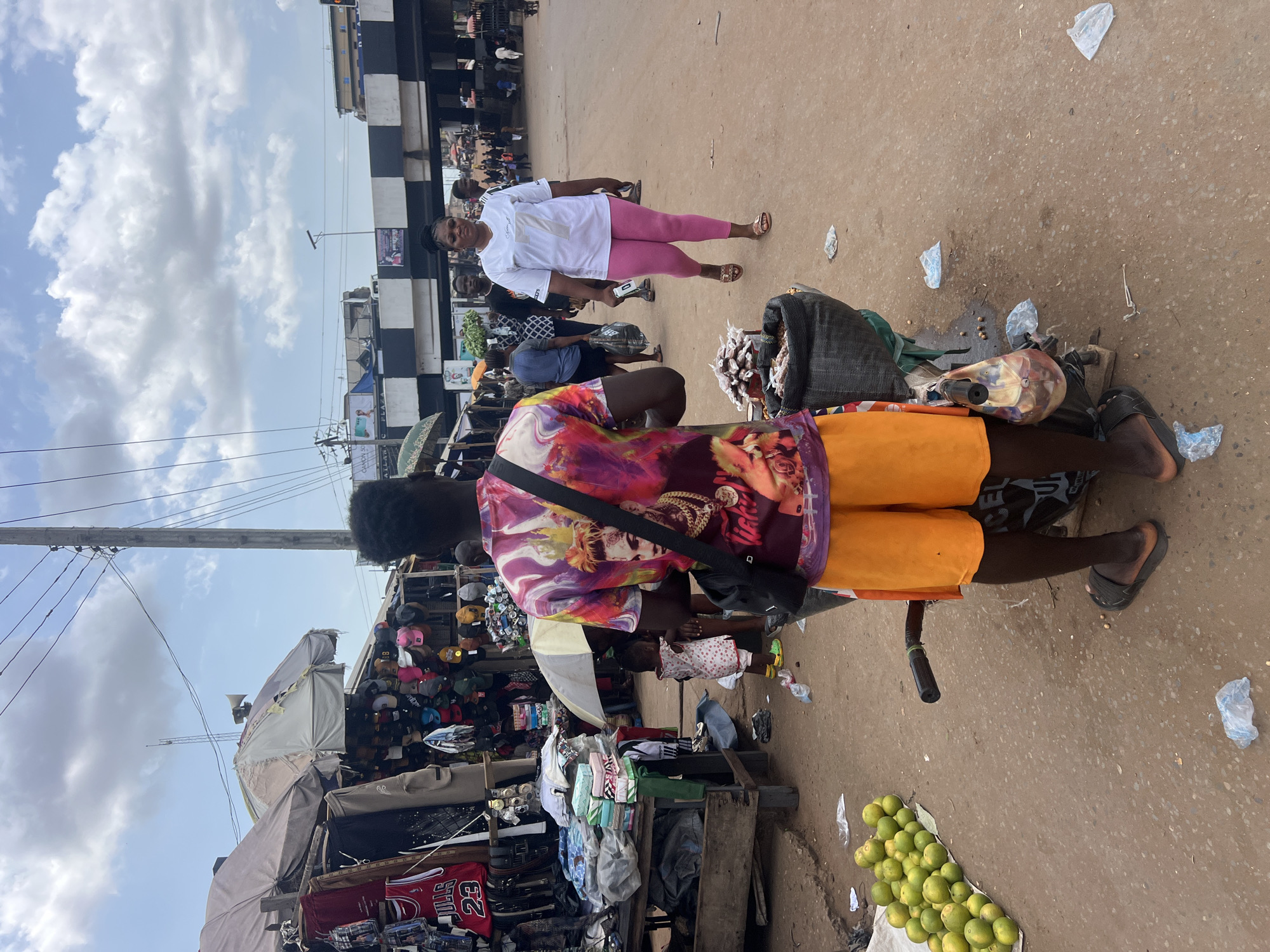 A daytime outdoor scene depicts a bustling street market in Unknown, Unknown. The market area is crowded with stalls and pedestrians. A man with dark skin and an afro, wearing an orange pair of shorts and a multi-colored t-shirt with a visible picture of a person, is standing on a bicycle. He has a black bag slung across his body. Next to him on the right is a pile of green citrus fruits on a white cloth. Behind the man, a woman with fair skin is walking towards the camera, wearing a white shirt and pink leggings. There are many stalls in the background, filled with various goods such as clothing, hats, and other items. The stalls are covered with umbrellas for shade. The ground is uneven with some litter, and there is a building with a black and white checkered pattern in the distance. The sky is partly cloudy.
