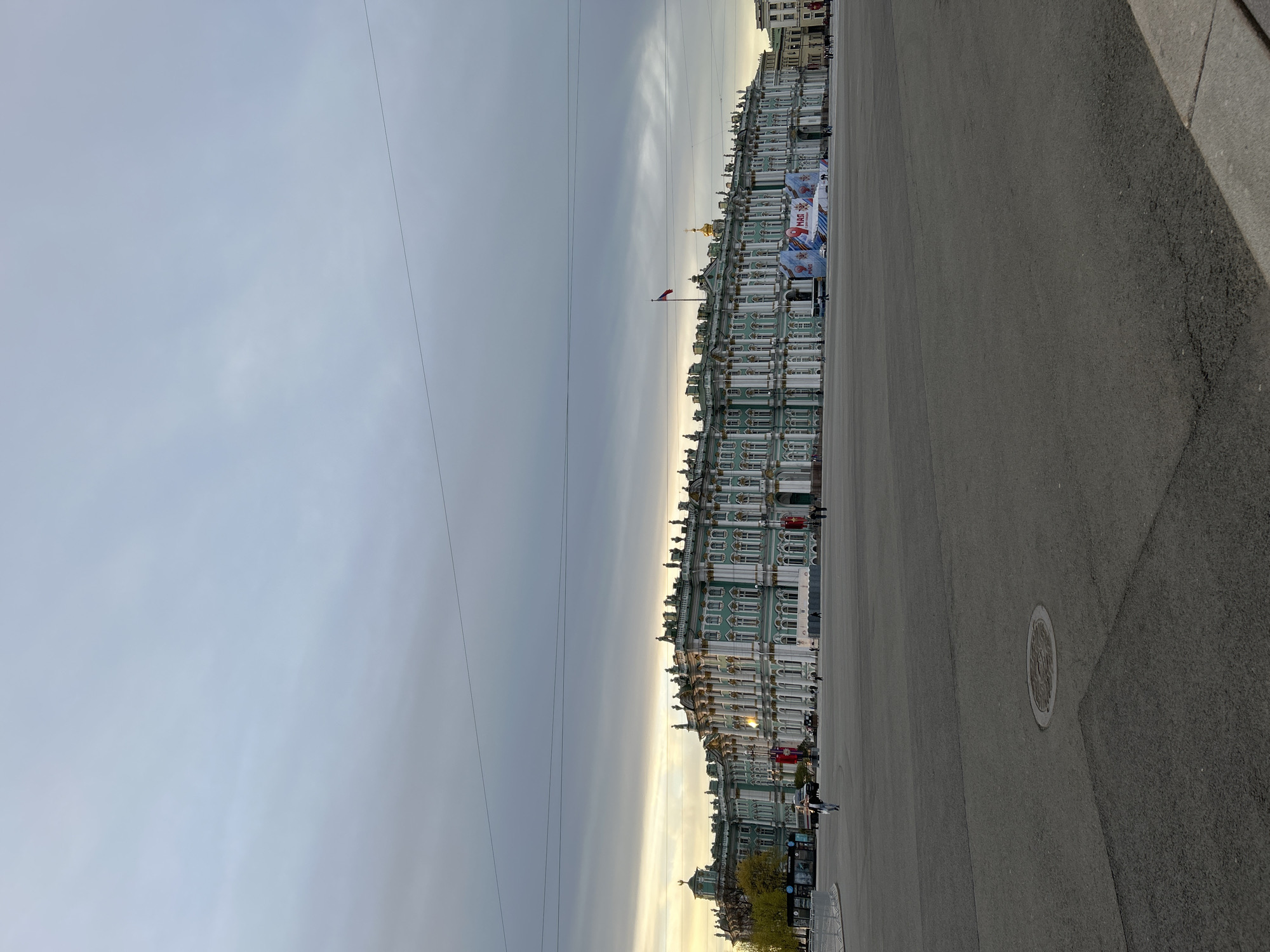 The image depicts Palace Square in Saint Petersburg, Russia. The Winter Palace is visible on the right side of the frame. The square itself is paved with asphalt, and there is a utility access cover in the foreground. Power lines are visible in the sky. Several banners celebrating May 9 are strung between the columns. A Russian flag flies on the roof of the Winter Palace. A few pedestrians are visible in the distance. The sky is overcast.