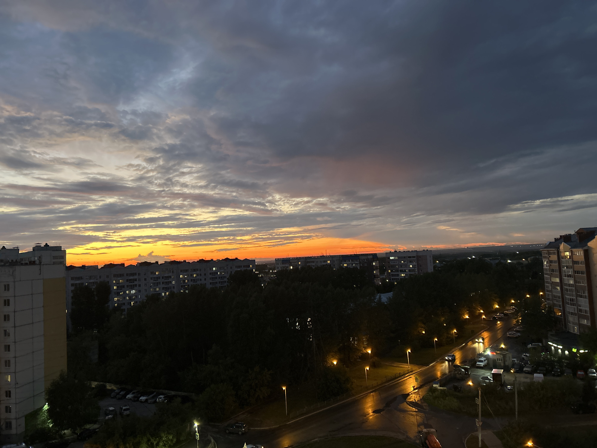The image captures a twilight scene over a residential area in Tomsk, Russia. The sky is filled with dramatic clouds, transitioning from a vibrant orange glow near the horizon to dark, heavy clouds overhead. In the foreground, several apartment buildings are visible, some with lit windows, suggesting the early evening. Dense trees line the streets, and streetlights cast a warm glow on the wet asphalt, indicating recent rain. Cars are parked on the street and in a parking lot, and a few are visible on the road. The overall atmosphere is one of a city settling in for the night under a dynamic sky.