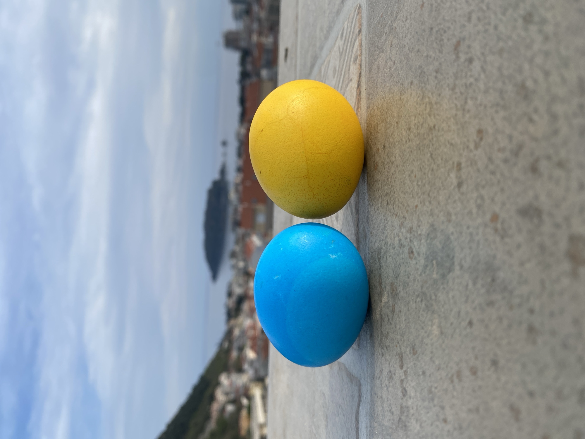 Two spherical objects, one yellow and one blue, are arranged vertically on a grey, weathered concrete surface. In the background, a blurred cityscape stretches out, suggesting the city of Budva, Montenegro, with its characteristic terracotta rooftops and stone buildings. Beyond the city, a body of water and distant mountains can be seen under a partly cloudy sky. The angle of the photograph suggests a high vantage point. There are no people visible in the scene.