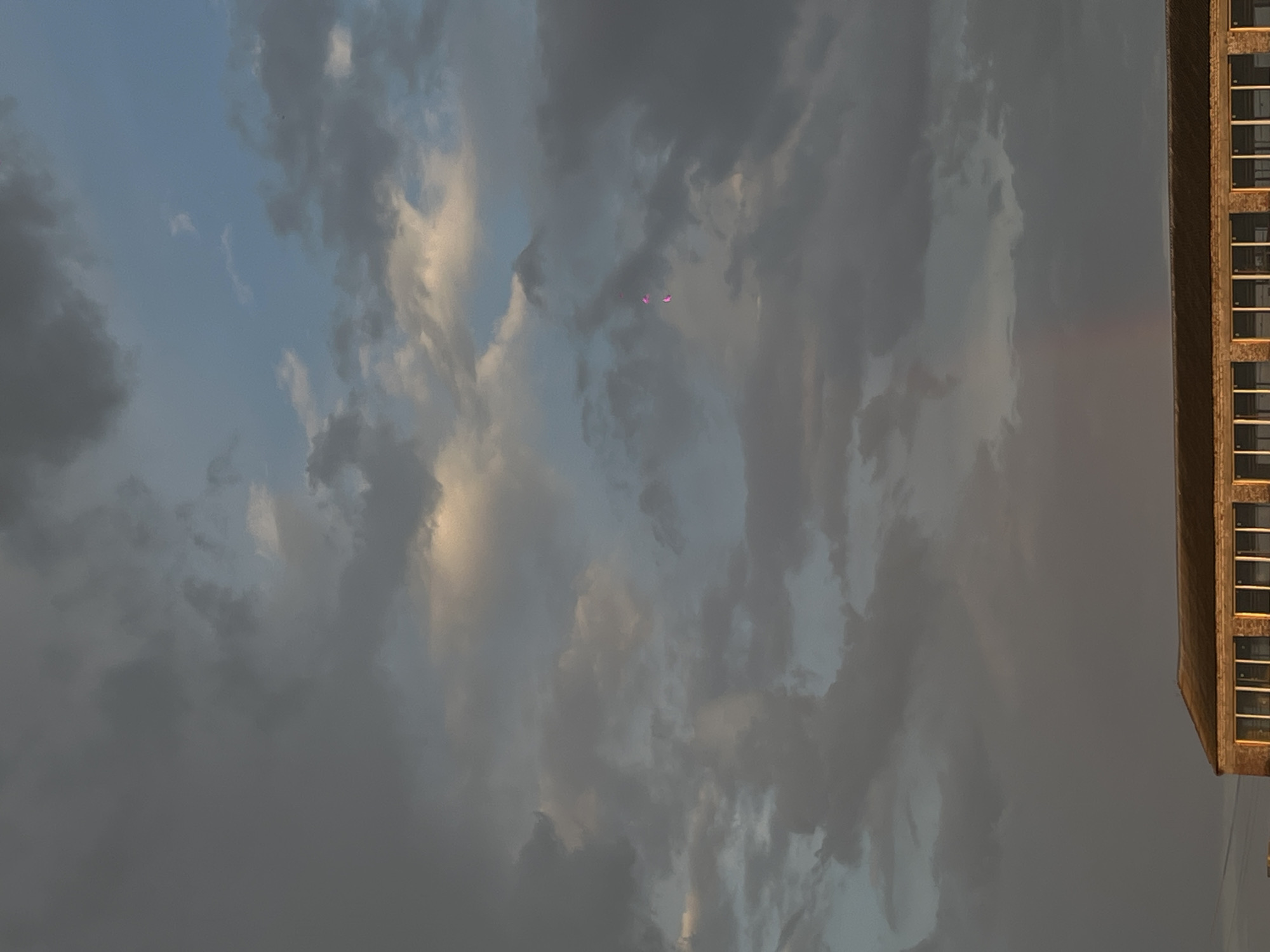 The image shows a cloudy sky with a building visible on the right side. The building appears to be a multi-story structure with multiple windows. There are three objects with pink colored reflection in the sky. The image does not contain identifiable people. Location of the image is not available.
