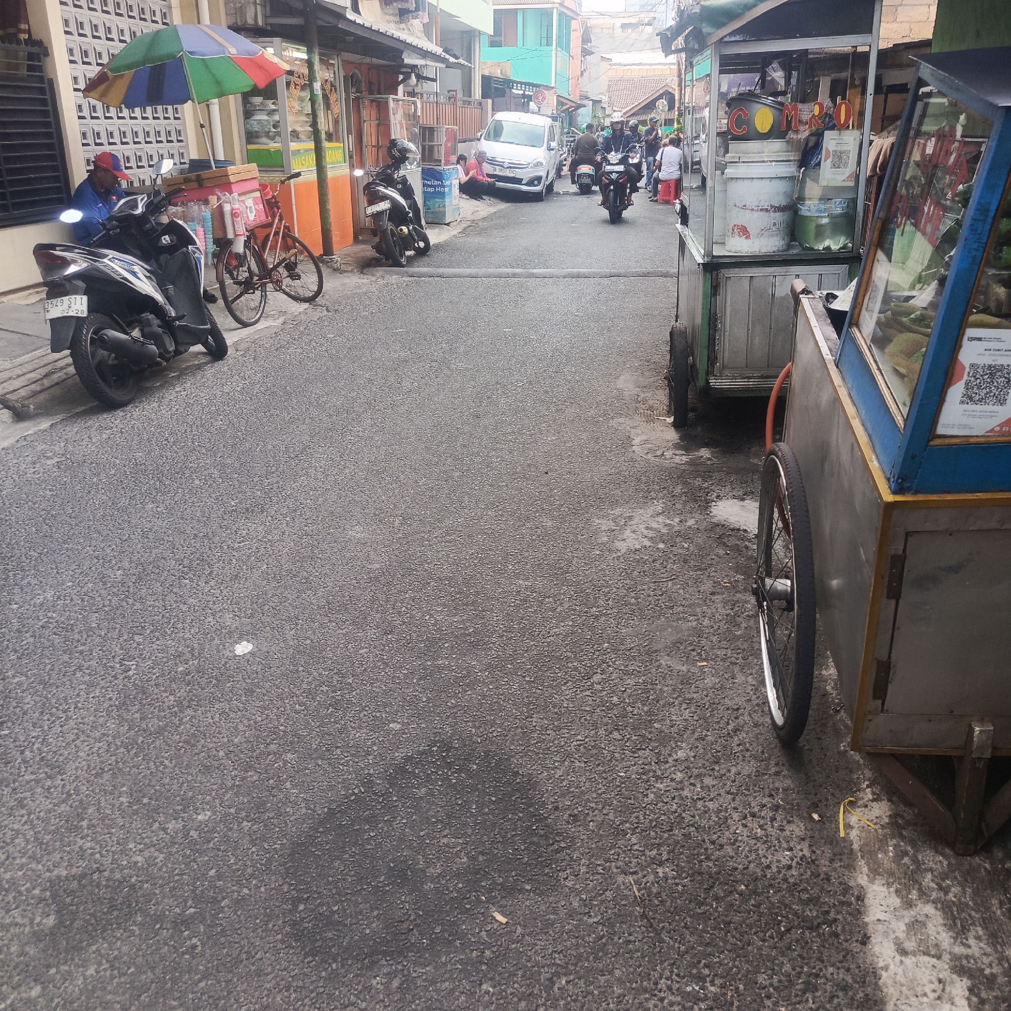 In the city of South Jakarta, Indonesia, an outdoor scene depicts a narrow asphalt street lined with low-rise buildings and various carts. To the left, a person wearing a blue jacket and a red and blue baseball cap sits on a motorcycle parked alongside a bicycle. A colorful umbrella is set up over a cart stocked with drinks. Further down the street, a white van is parked, and several other motorcycles are visible, some with riders. A food cart with a glass display case and a QR code sticker is prominently featured in the foreground on the right. The environment suggests a bustling, possibly commercial area with street vendors and local transportation.