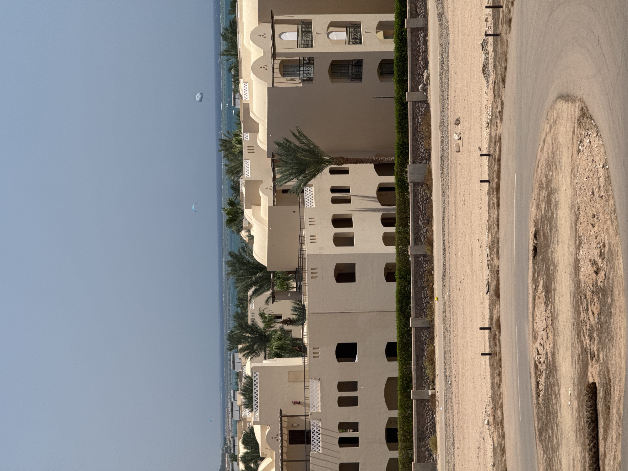 An exterior, eye-level, vertical shot captures a resort complex on a bright, clear day in an arid environment, likely near a body of water. The resort buildings are low-rise, constructed with light-colored stucco and featuring rounded archways and small balconies with decorative railings. Palm trees are interspersed throughout the property. In the background, a thin strip of ocean is visible, with two distant paragliders visible against the hazy blue sky. The foreground is dominated by a dusty, unpaved area with a paved road curving through it. The overall scene suggests a warm climate and a leisure-focused destination.