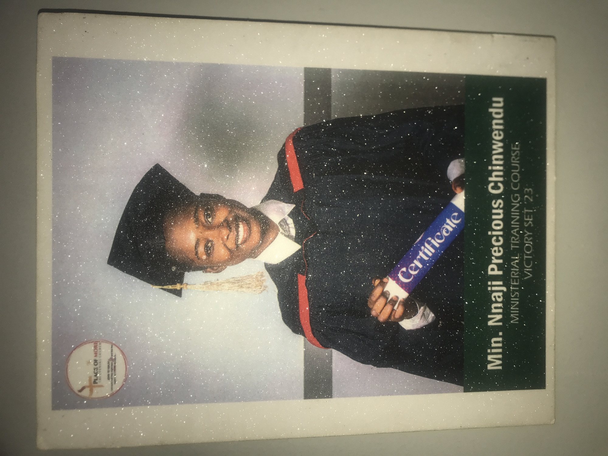 A vibrant portrait captures a young woman, Min. Nnaji Precious Chinwendu, smiling broadly in graduation attire, celebrating her completion of a ministerial training course in Rafin Par, Nigeria. The image is rotated 90 degrees counter-clockwise. She wears a black graduation gown with red trim and a matching mortarboard. In her hands, she holds a rolled-up document clearly labeled "Certificate." Her joyful expression conveys a strong sense of achievement and happiness. The background is a light, diffuse setting, possibly a studio, with subtle vertical elements. A prominent feature of the photograph is a glittery, sparkly effect applied uniformly across the entire image, adding a celebratory feel. In the bottom left corner, a circular emblem is visible, containing text that includes "PLACE OF MORE" and "THRIVING CHURCH," along with "REV. DR. SEGUN ADEKANBI" below it. Vertically oriented text on the right side of the image further identifies her as "Min. Nnaji Precious Chinwendu" and details her accomplishment: "MINISTERIAL TRAINING COURSE. VICTORY SET 23."