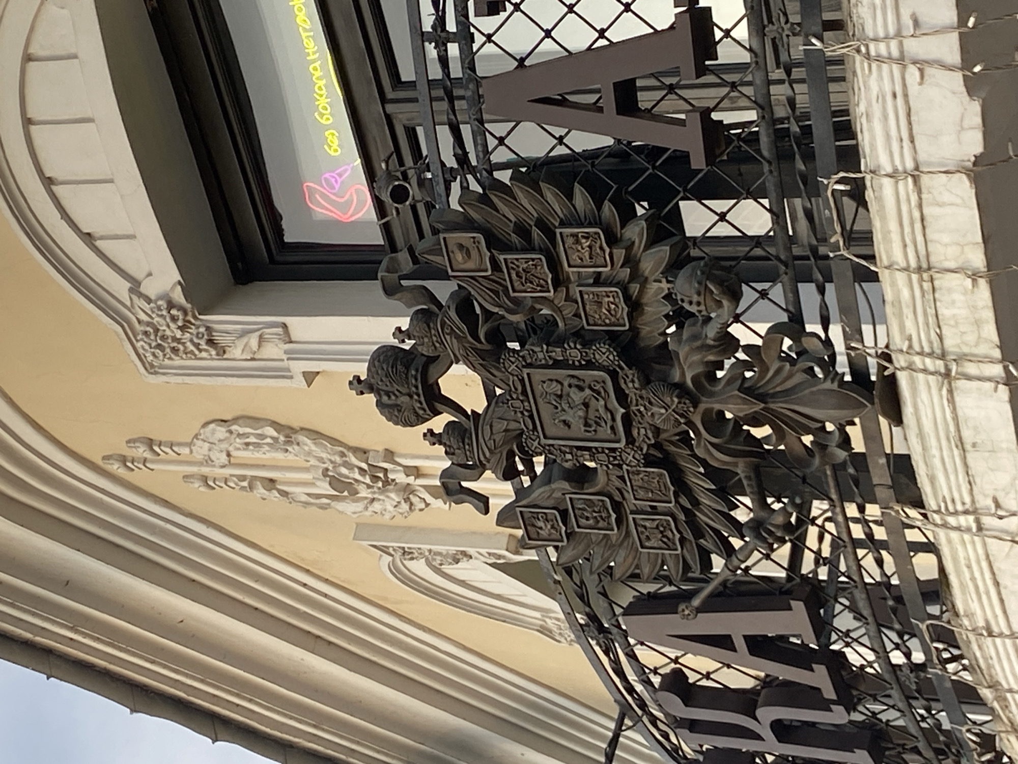 The image depicts an ornate, metallic coat of arms with crowns and detailed emblems affixed to a structure, possibly a building facade. Behind the coat of arms is a metal framework with the letters "HARR" visible. The architecture suggests a European style, possibly from the 19th century. In the background, a neon sign with Cyrillic text and a hand graphic is visible through a window. A cream-colored building with layered molding is apparent. Given the architectural style and the presence of Cyrillic text, the location is likely in Russia, and based on the prevalence of similar architecture, Kazan is a possible location.
