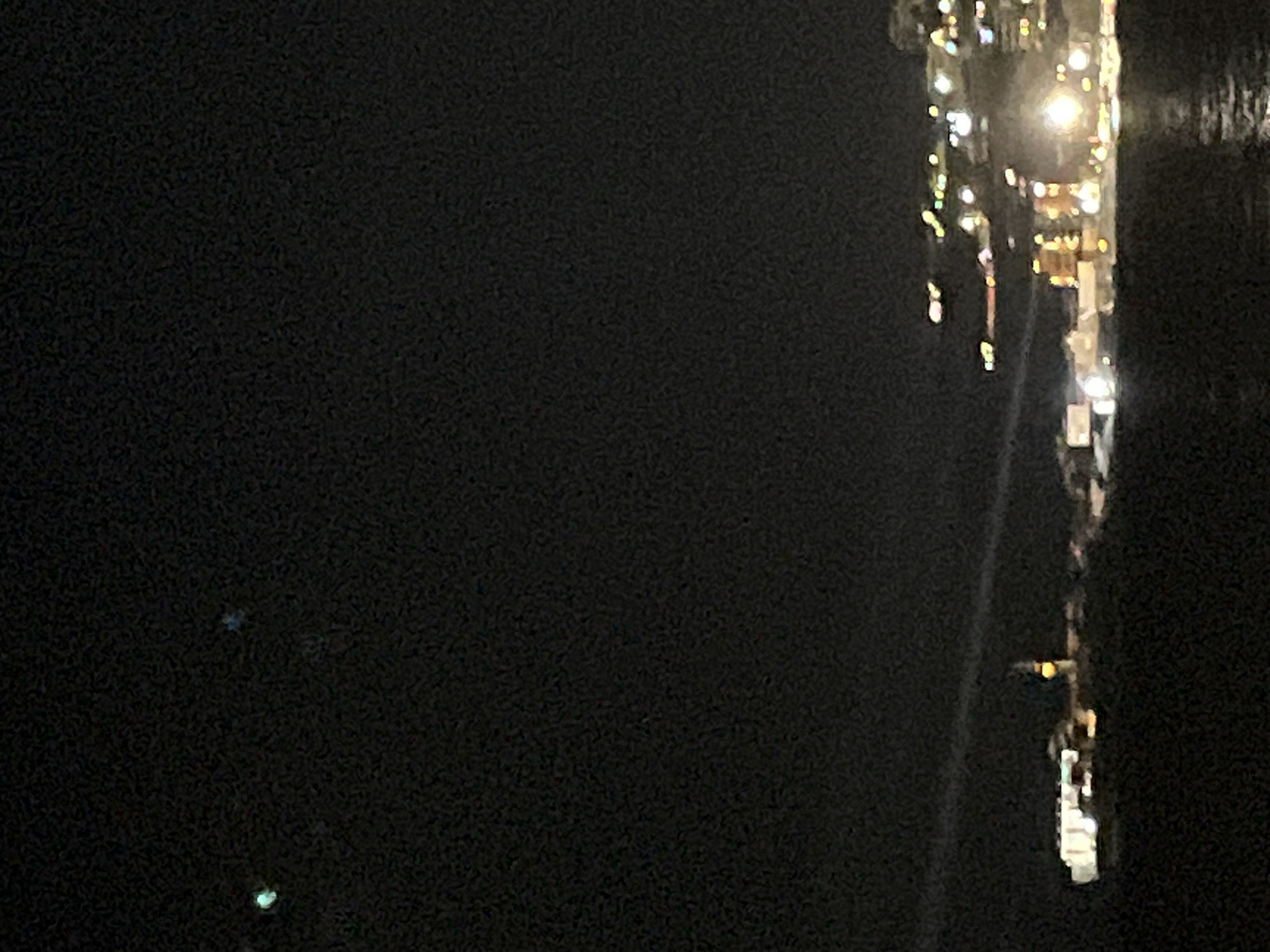 The image presents a nighttime panoramic view of Budva, Montenegro. The upper two-thirds of the frame consists of a dark, grainy sky, exhibiting a faint green-blue point light on the far left and a fainter blue-grey point light slightly to its right. A faint, light-colored linear streak extends horizontally across the midground, above the distant landmasses. The lower third of the image shows a dark body of water, likely the Adriatic Sea, with blurry reflections of artificial lights.
On the left side of the midground, a distinct landmass, identified as Sveti Nikola Island, is partially illuminated. Multiple white and yellow lights mark structures on this island, with a prominent, tall structure featuring a bright orange-yellow light at its apex, characteristic of a lighthouse. To the right of the frame, the main city of Budva is visible as a densely populated area ascending from the coastline. This landmass is illuminated by numerous warm white and yellow artificial lights, indicating buildings, roads, and urban infrastructure. No discernible individuals, specific actions, or direct interactions are visible within the scene.