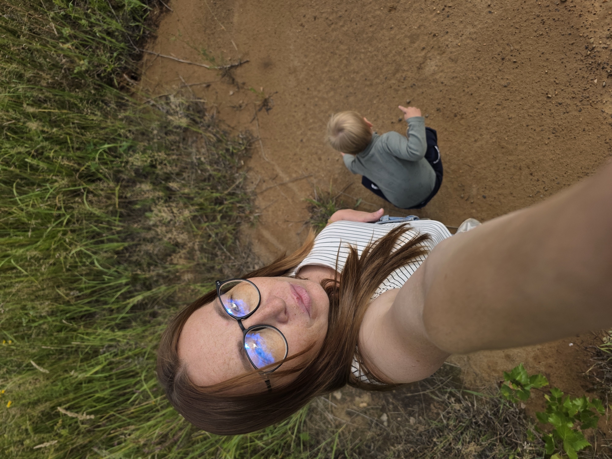 In this overhead, full-body selfie, a woman with auburn hair and freckles is lying on her back in the grass, holding the camera above her. She is wearing glasses with reflective blue lenses and a black and white striped shirt. Her expression is relaxed and content. To her right, a young boy with blonde hair is sitting on a dirt path, pointing towards something out of frame. He is wearing a grey long-sleeved shirt and dark shorts. The scene appears to be outdoors, with lush green grass on the left and a dry, sandy path on the right. The lighting suggests it is daytime, possibly overcast given the soft shadows. The environment looks natural and uncultivated, possibly a park or a rural area. The overall mood is one of casual enjoyment and a shared moment between the woman and the child. There is no visible text in the image.