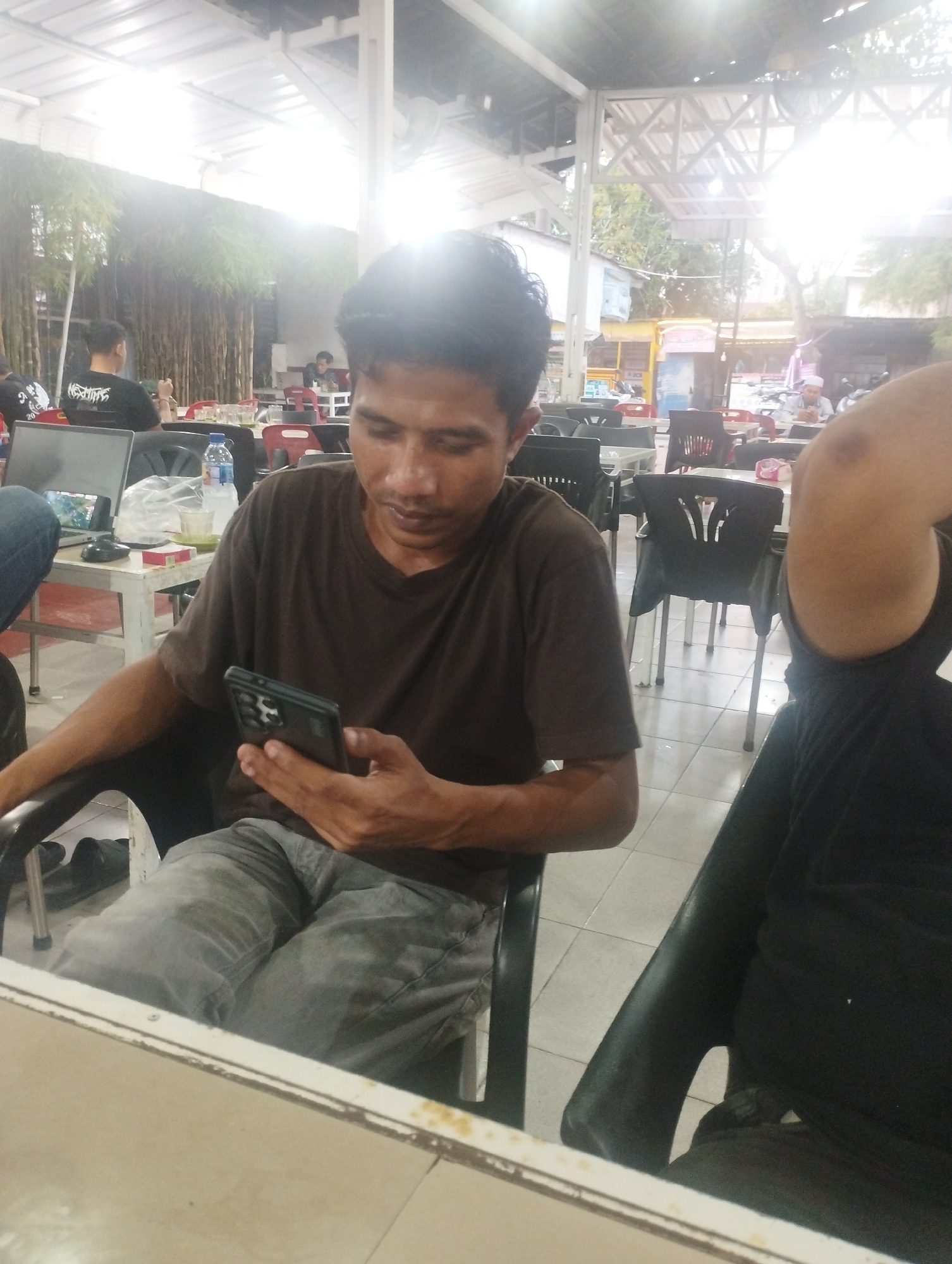 The image shows a man sitting at a table in an open-air restaurant. He is holding a black smartphone in his hands and looking at the screen. He is wearing a brown t-shirt and gray jeans. Another person wearing a black shirt is partially visible on the right side of the frame. In the background, there are other people sitting at tables, a laptop, water bottles, and cigarettes on a table. The location is Lhokseumawe, Indonesia. The building has a partial roof with a visible roof structure. Vegetation can be seen outside.