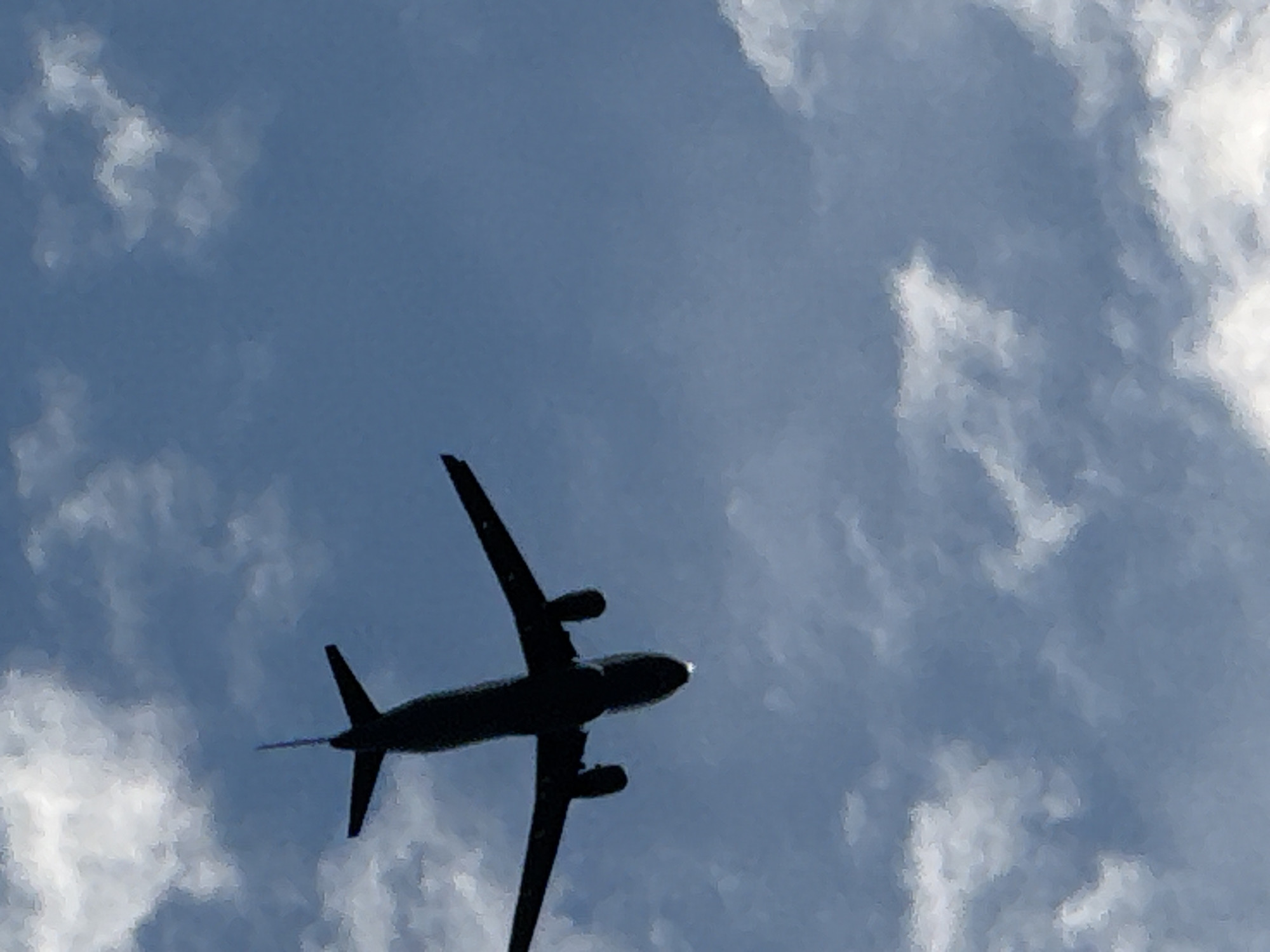 A silhouette of a commercial airplane is captured from below against a blue sky filled with white clouds. The airplane is angled slightly, with its wings spread and engines visible. The image does not provide any visual information that would allow for the identification of the city of Budva, Montenegro.