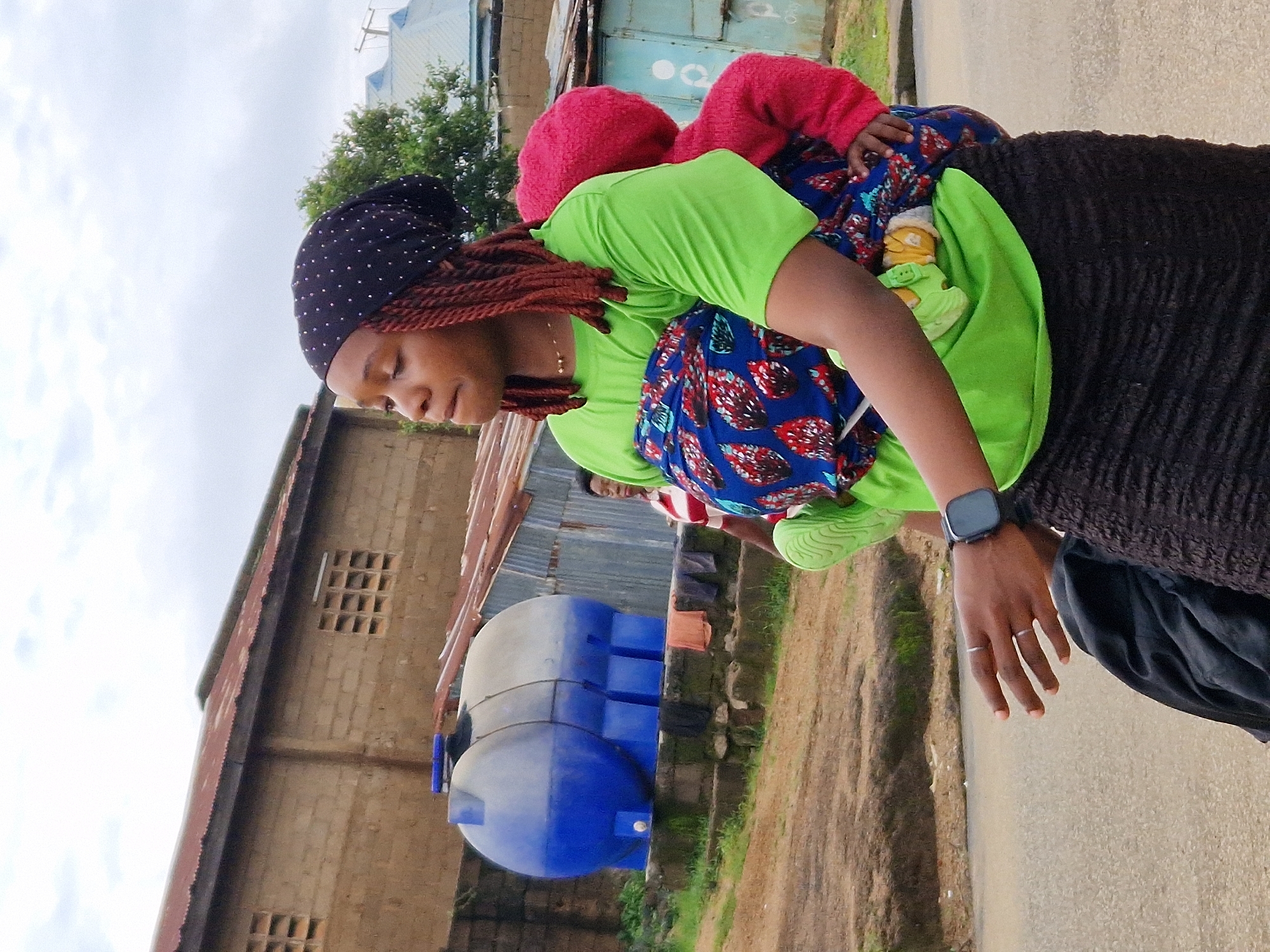 A medium shot captures a woman walking outdoors, carrying a baby wrapped in a colorful cloth sling. The woman, wearing a lime green t-shirt, a black headscarf adorned with white dots, and a dark, textured skirt, has her hair in long, reddish-brown braids. A smartwatch is visible on her left wrist, and she wears two rings on her left hand. The baby, dressed in a pink hooded garment, is secured to her back. In the background, a large blue water tank stands near a brick building with a corrugated metal roof. Another person in a striped shirt is partially visible in the middle ground, standing near the water tank. The setting appears to be a dusty, outdoor area with a paved road in the foreground. The sky is overcast.